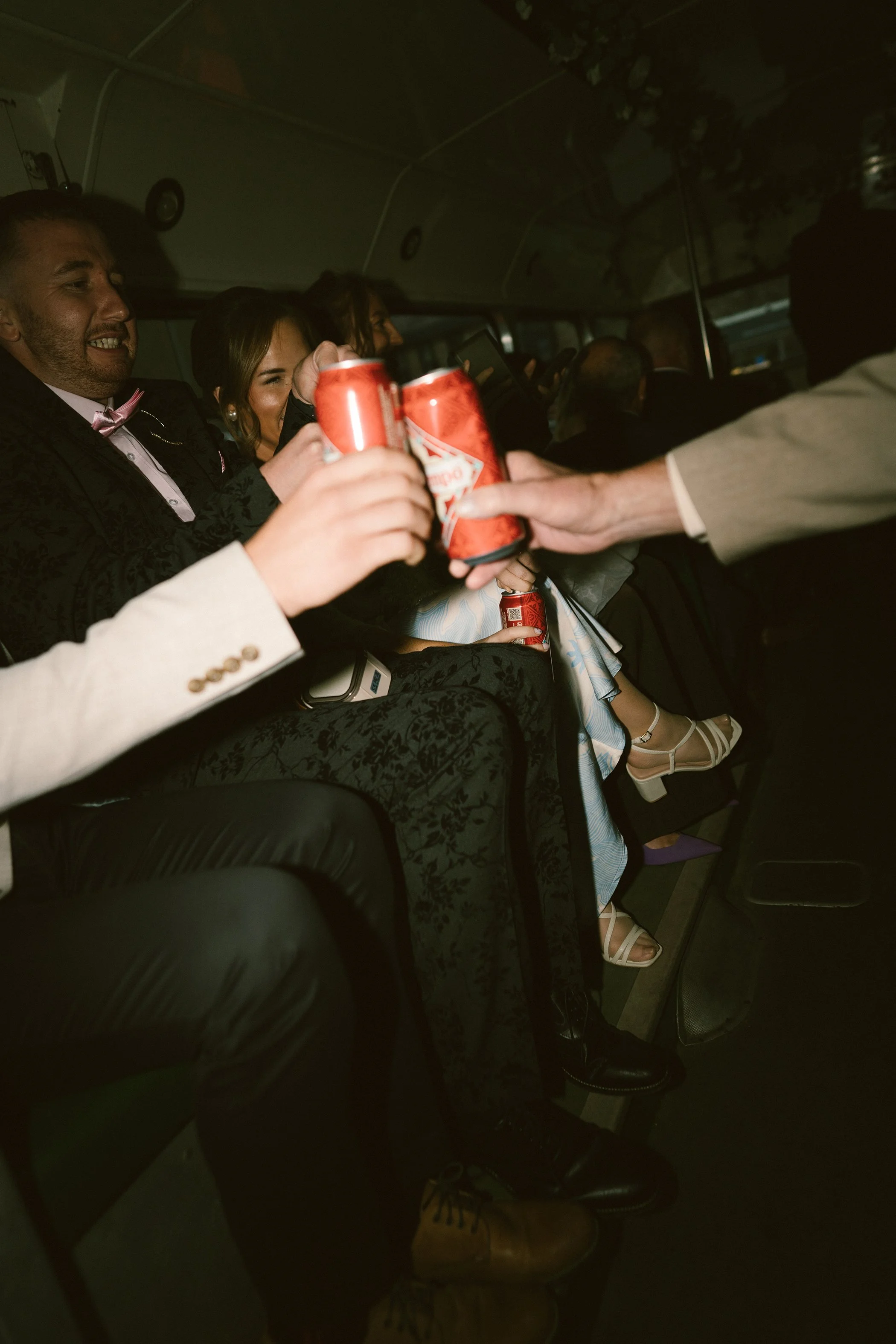 People sitting inside a vehicle, holding cans of beer, with some people smiling and looking towards the camera. The atmosphere appears to be lively and social.