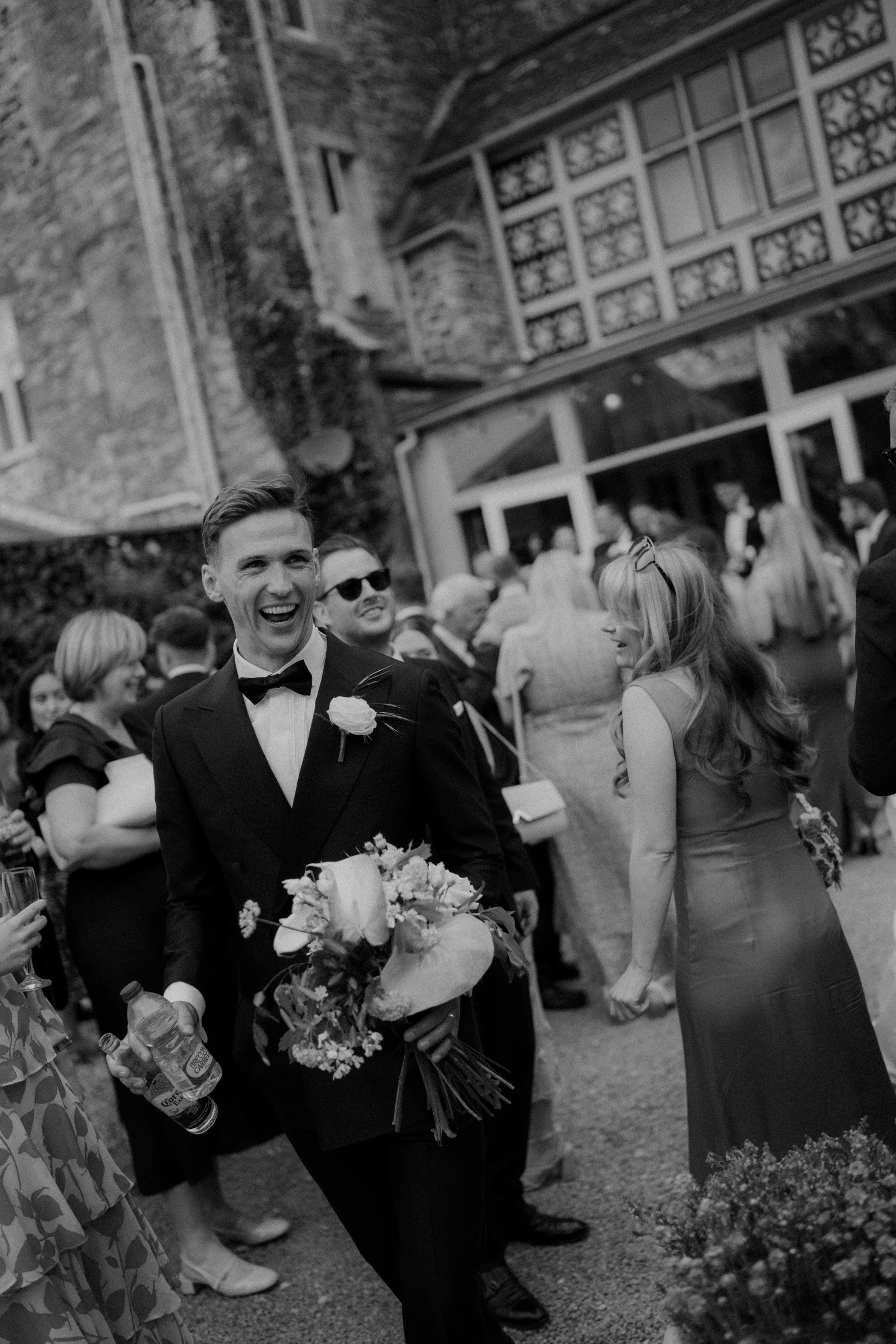 A black and white photo of a wedding celebration with a smiling groom in a tuxedo holding a bouquet, surrounded by guests outdoors, with a large building in the background.