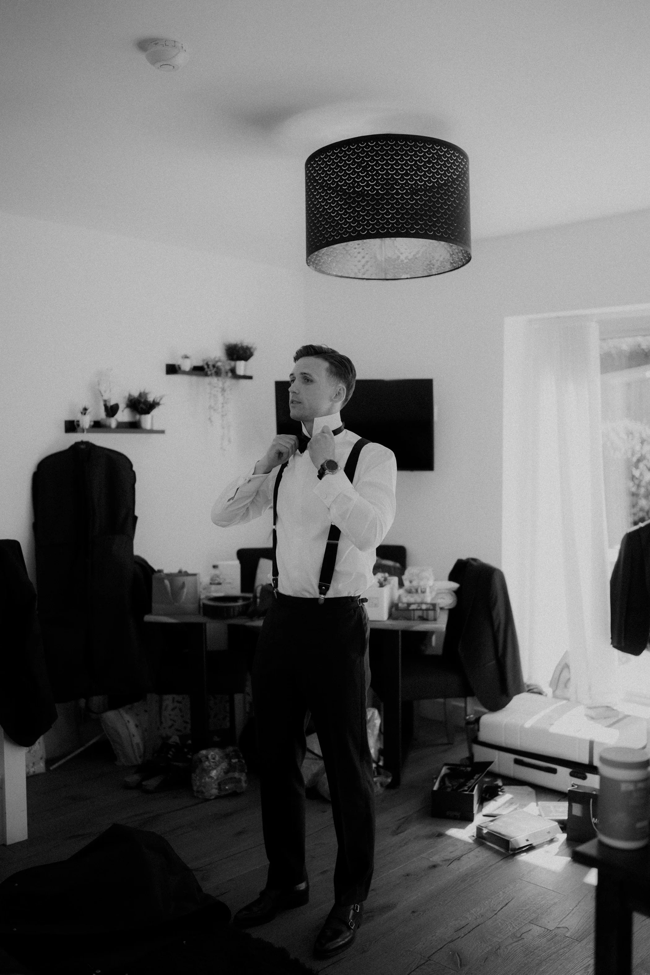 A man dressed in formal attire, adjusting his bow tie in a room with a modern decor, including shelves with plants, a television, and scattered items on furniture and floor.