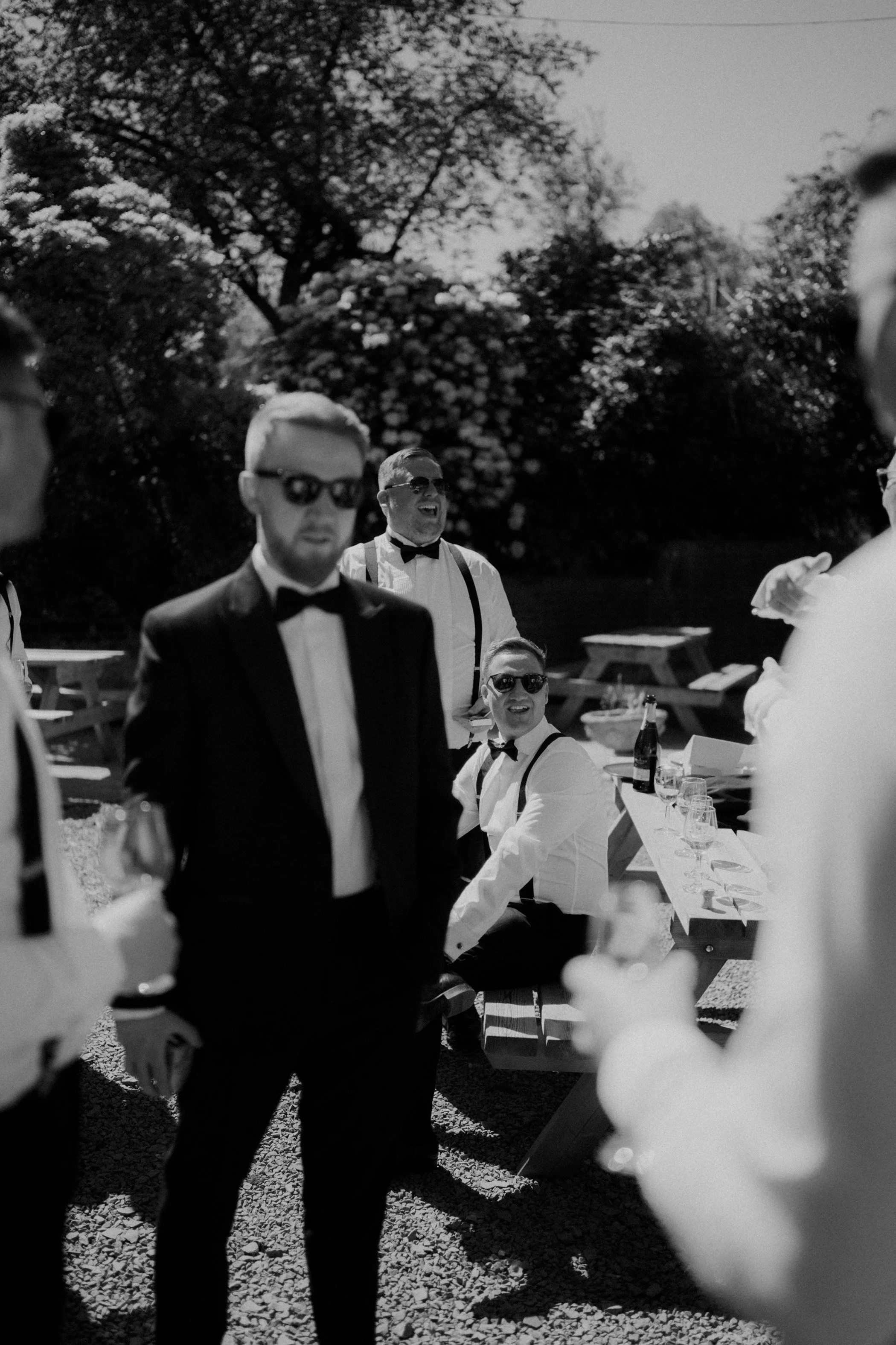 Black and white photo of men dressed in tuxedos and suspenders at an outdoor gathering with trees in the background, some wearing sunglasses, smiling and holding drinks.