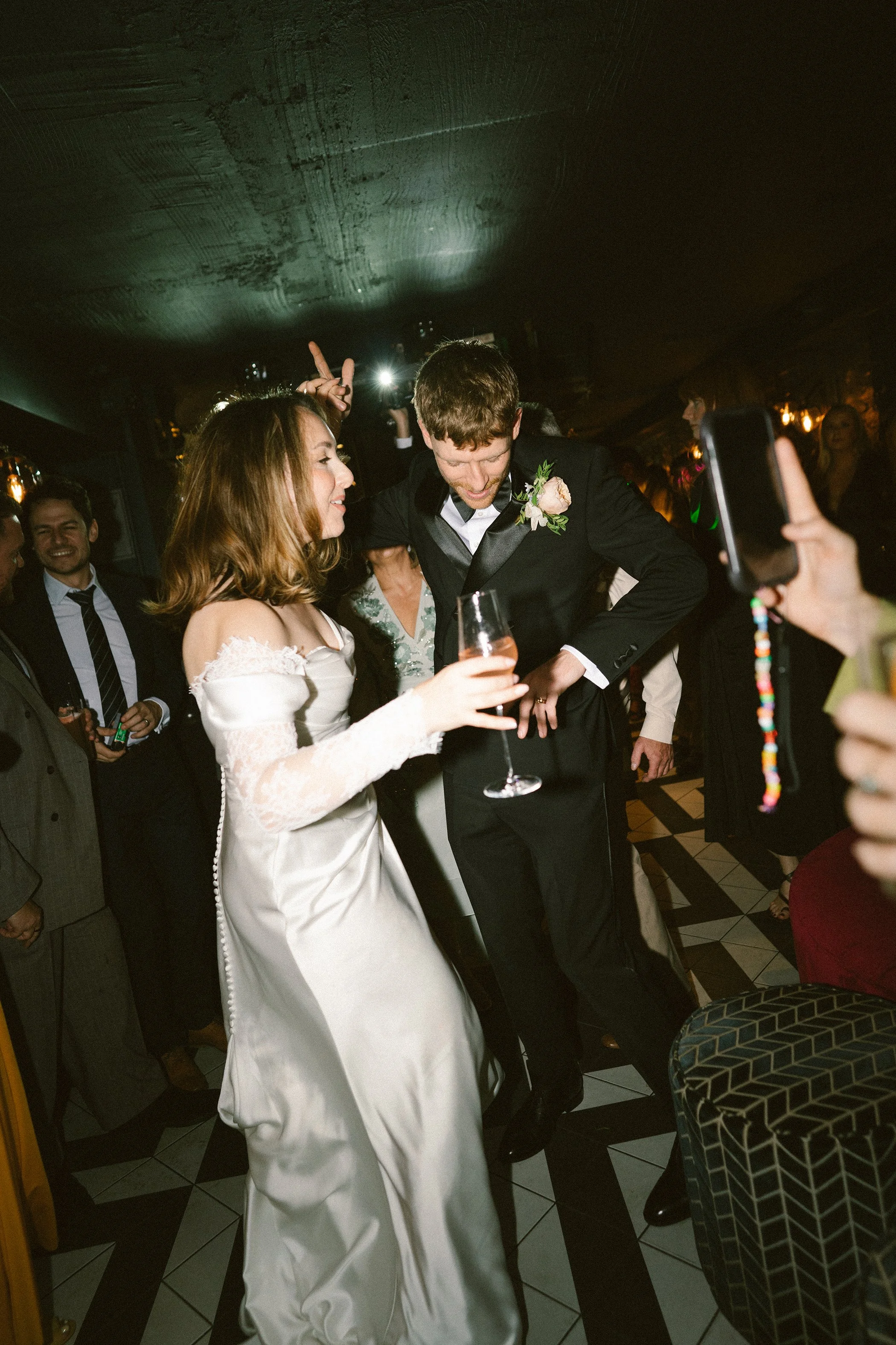 Bride and groom dancing at their wedding reception with guests around them, some taking photos or videos, in an indoor venue with dim lighting.