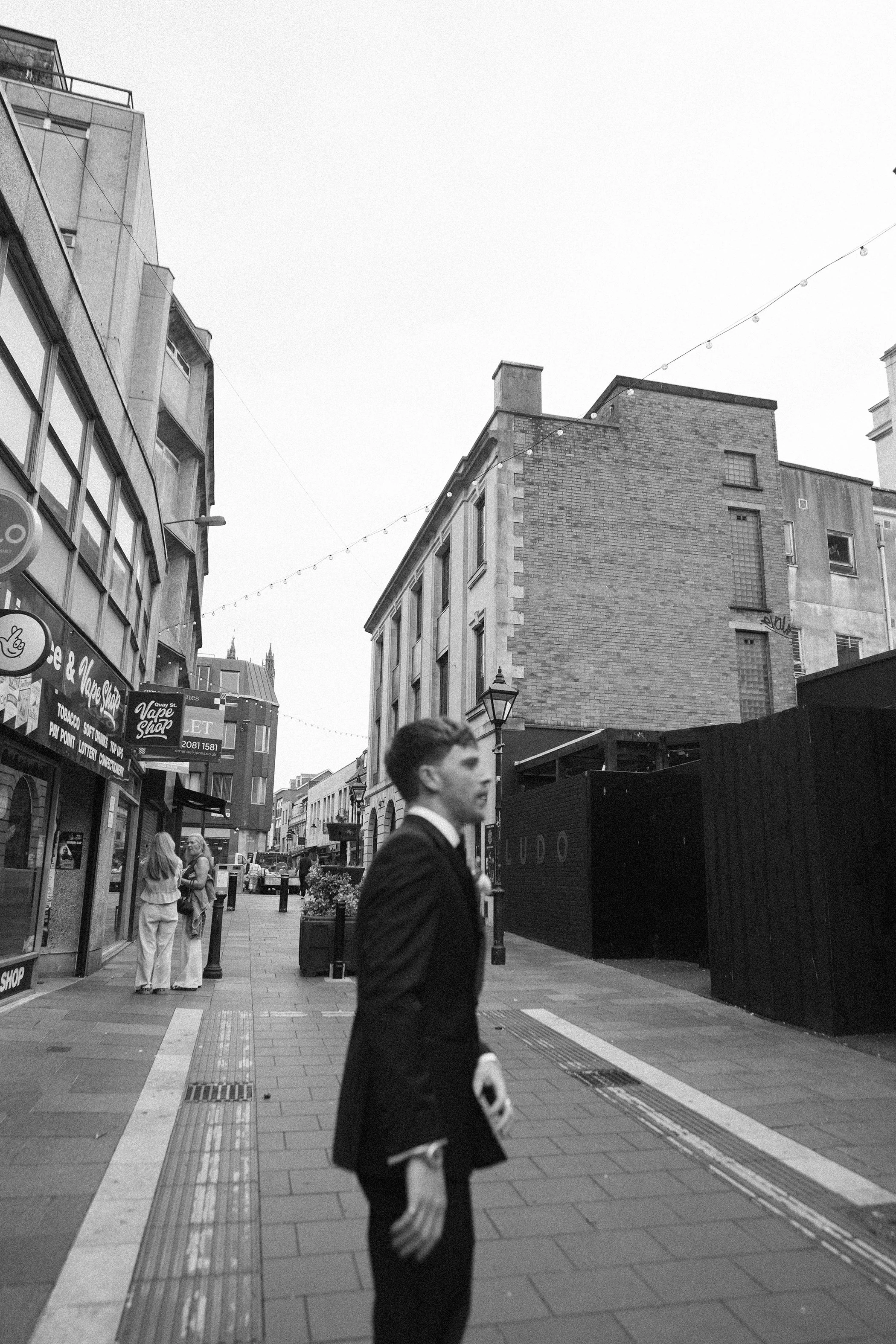 Black and white photo of city street with pedestrians, buildings, string lights, and a man in suit walking