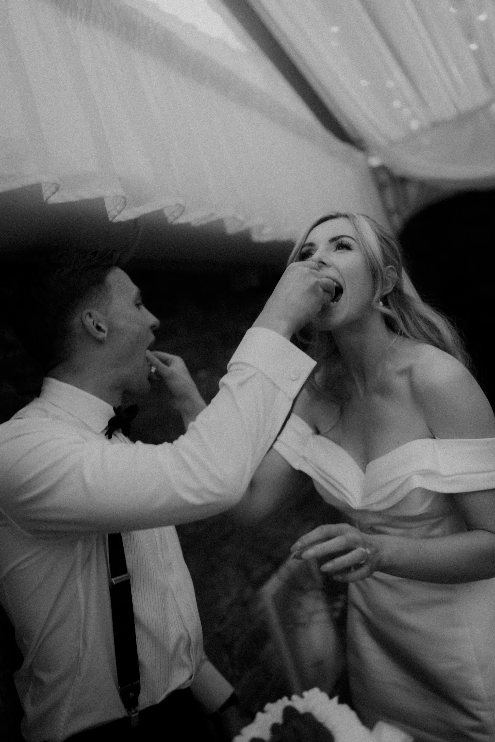 A black and white photo of a wedding reception where a groom is feeding a piece of cake to the bride during a cake-cutting ceremony.