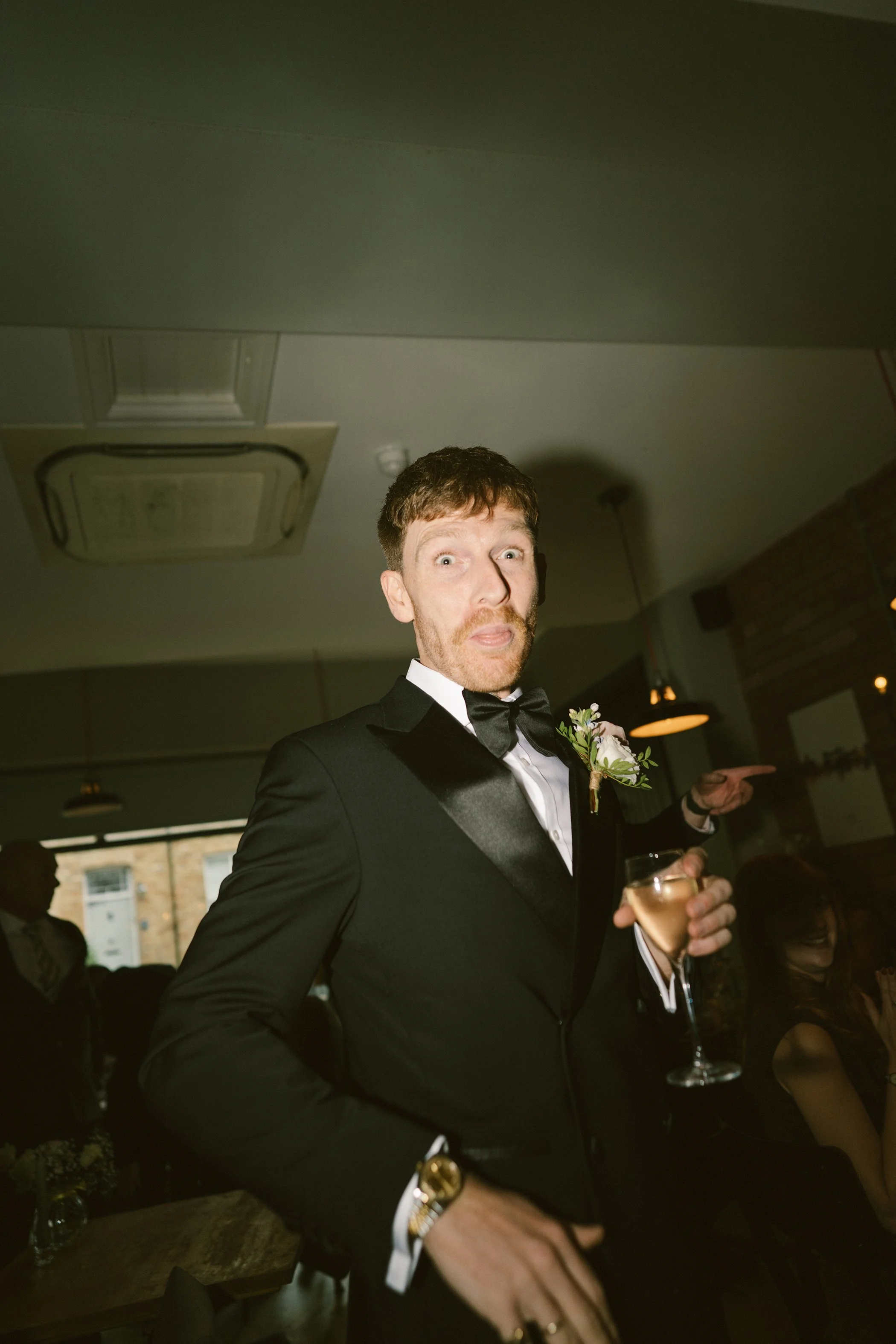 A man in a black tuxedo with a bow tie, holding a glass of champagne, at a social event or celebration, with a surprised or playful facial expression, in a dimly lit indoor setting.