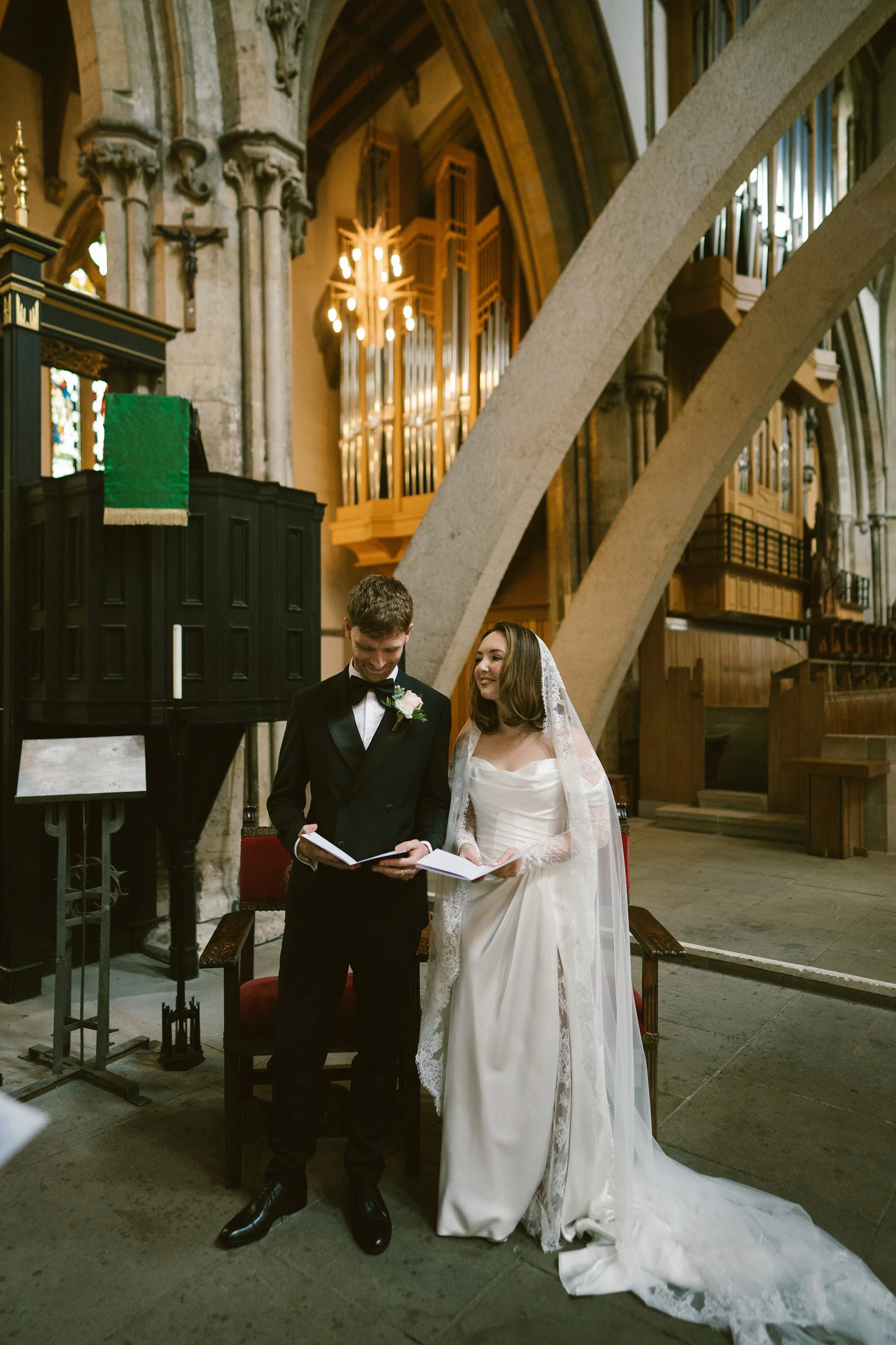 A bride and groom standing together in a church during their wedding ceremony. The groom is wearing a black tuxedo and the bride is wearing a white wedding gown with a veil. They are smiling and looking at each other, with a pianist and a church inte