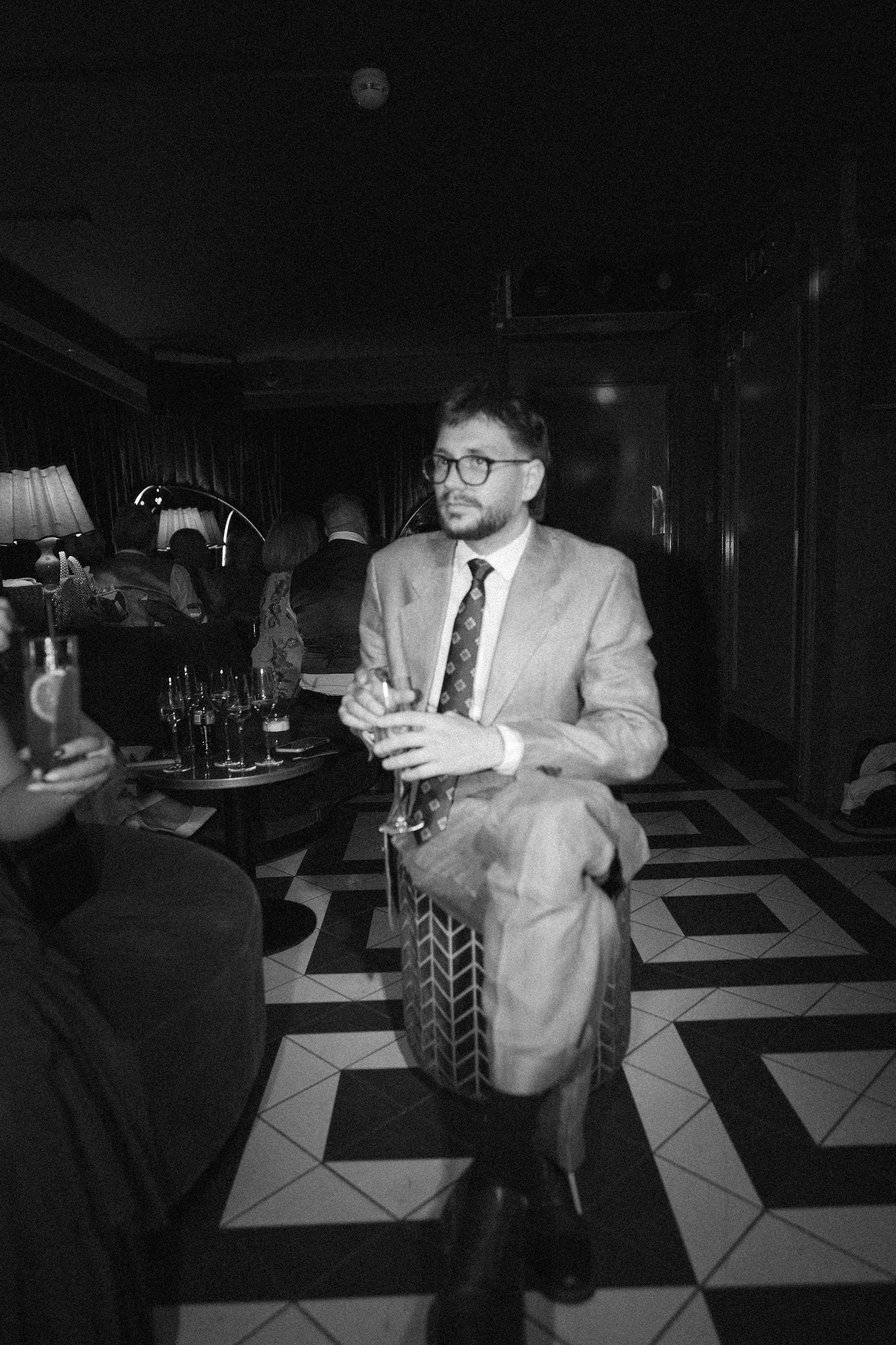 A man in a suit and tie, wearing glasses, holding a drink, standing in a dimly lit bar or lounge area with other people in the background, sitting at tables and talking.