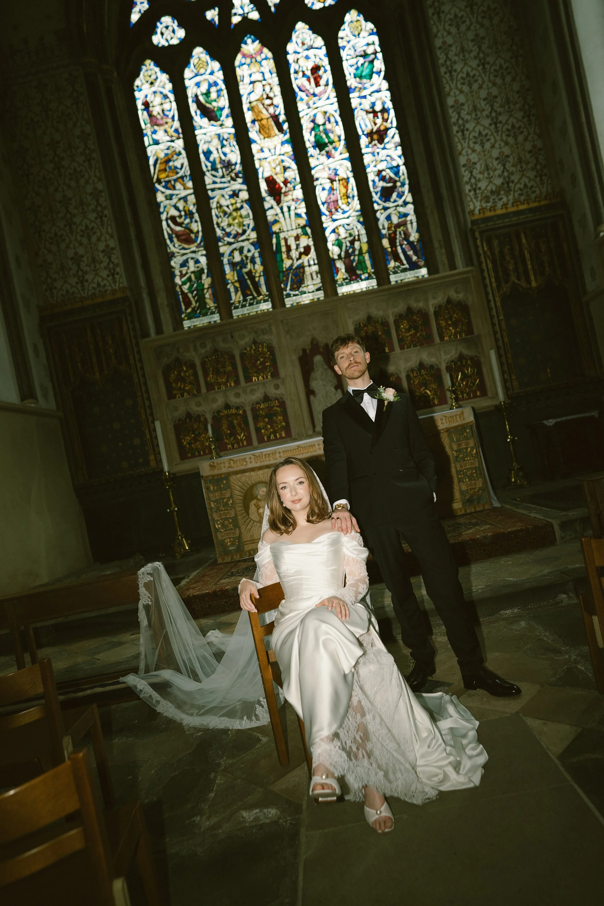 A bride in a white wedding dress and a groom in a black tuxedo with a bowtie inside a church with stained glass windows. The bride is sitting on a chair, and the groom is standing next to her, resting his hand on her head. The church interior feature