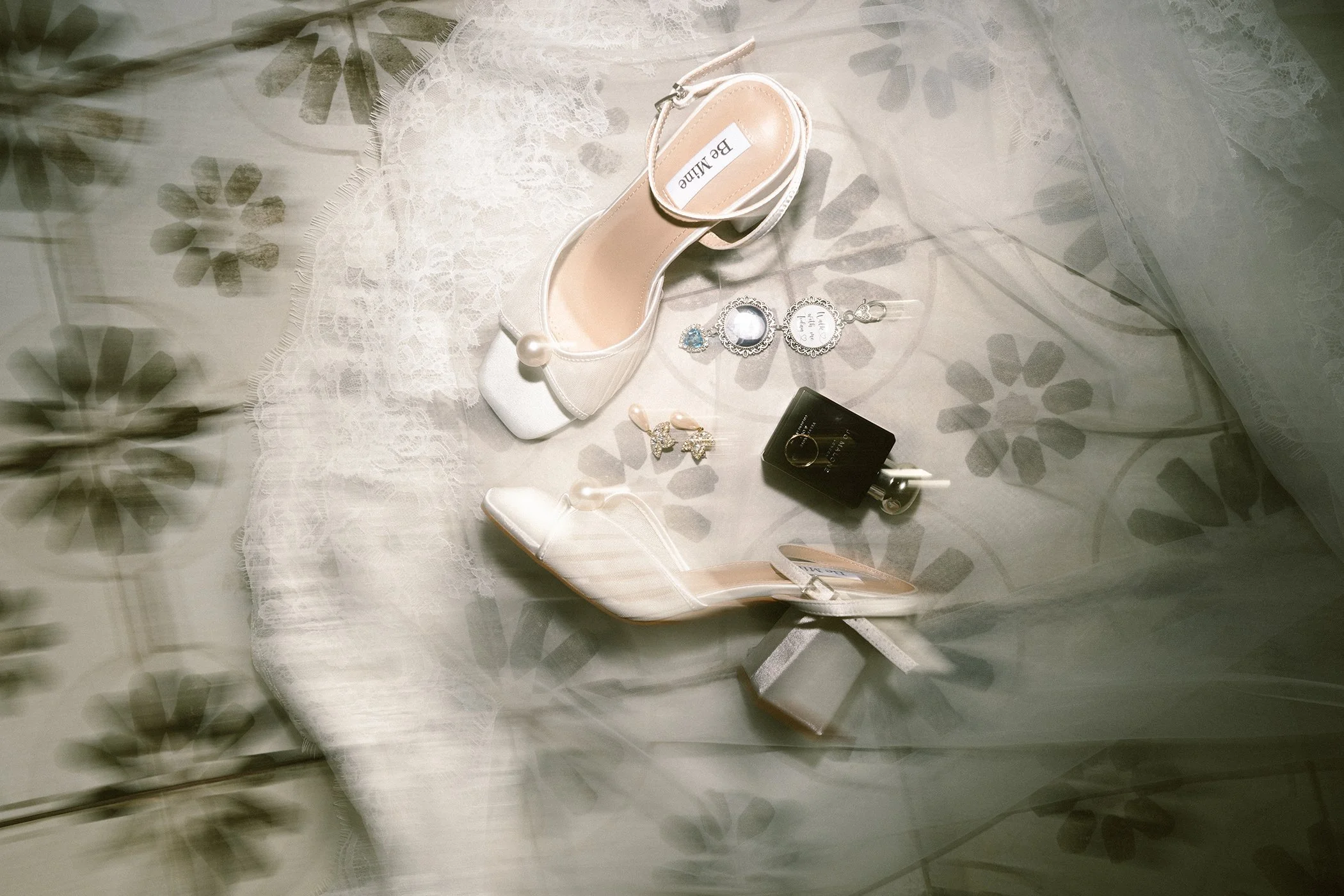 A white high heel shoe with a pearl decoration, a white shoe box labeled 'BE by BAILLE', a pair of pearl earrings, a ring, and a jewelry charm with a mirror and quote, all arranged on a lace-covered surface with floral shadow patterns.