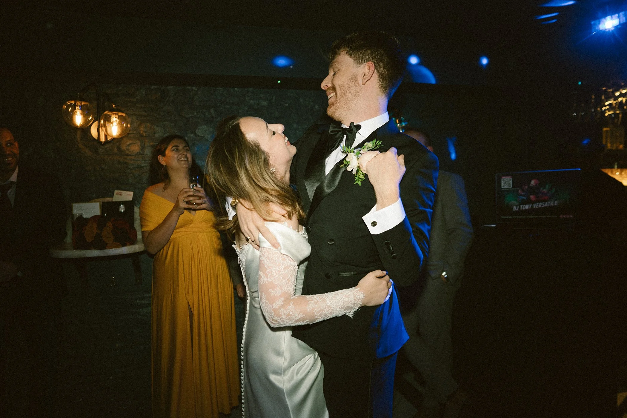 A man and woman dancing closely at a celebration, with people watching and smiling in the background, under warm lighting.