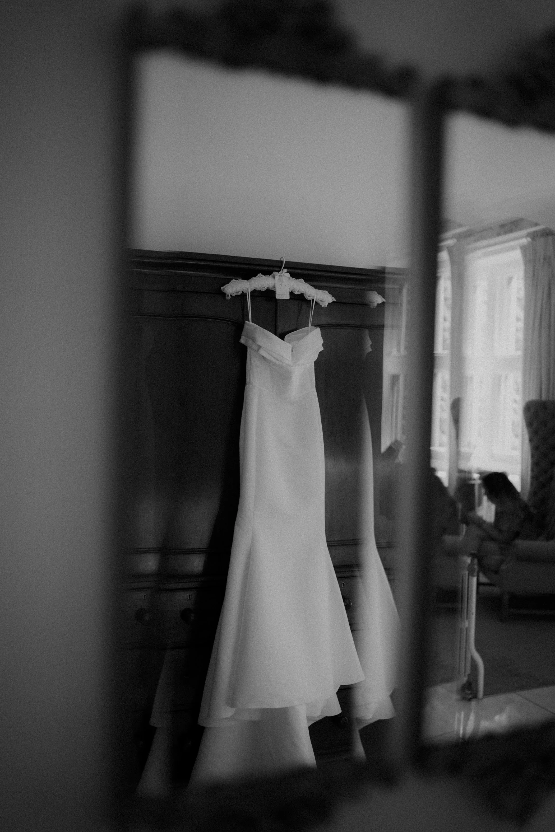 A white wedding dress hanging on a hanger in a bedroom, seen through a small opening or frame, with a person sitting in a chair reading a book in the background.