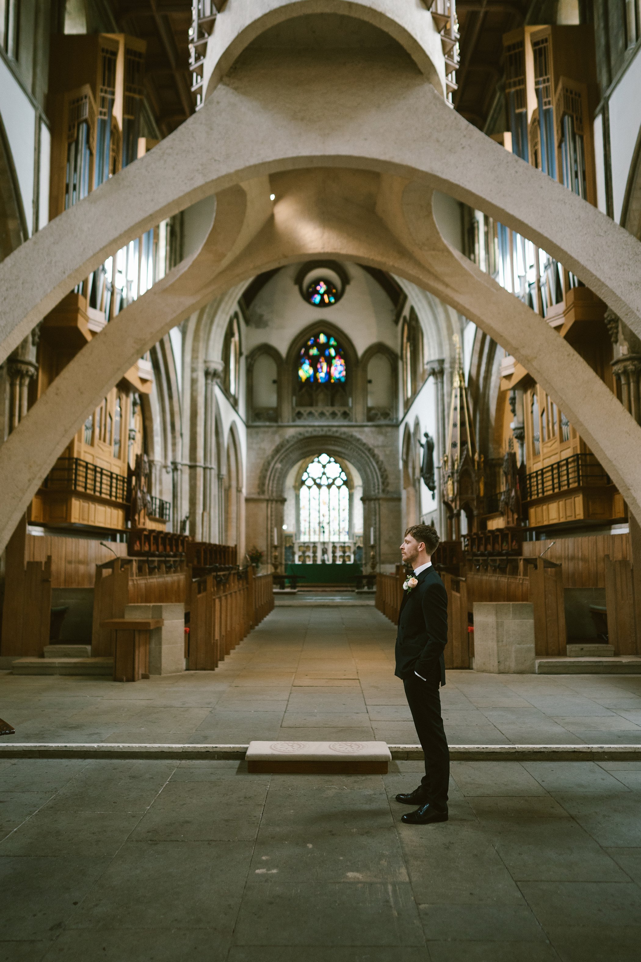 A man in a tuxedo standing inside a church with high vaulted ceilings, stained glass windows, wooden pews, and decorative arches.