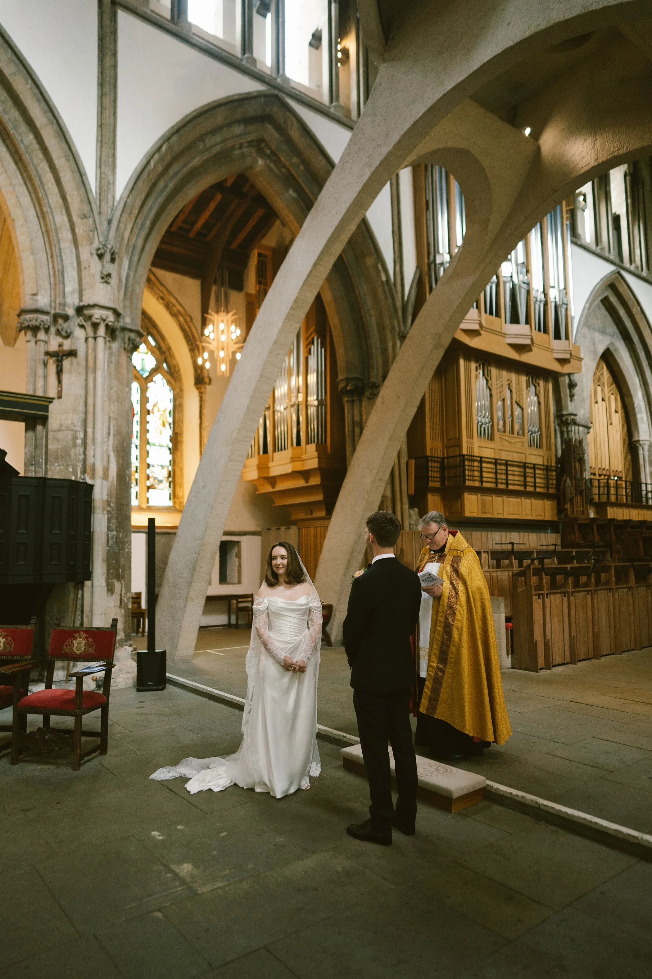 A bride and groom are standing before a priest during a wedding ceremony inside a historic church with high vaulted ceilings, stained glass windows, and large organ pipes. The bride is wearing a white wedding dress with lace sleeves, and the groom is
