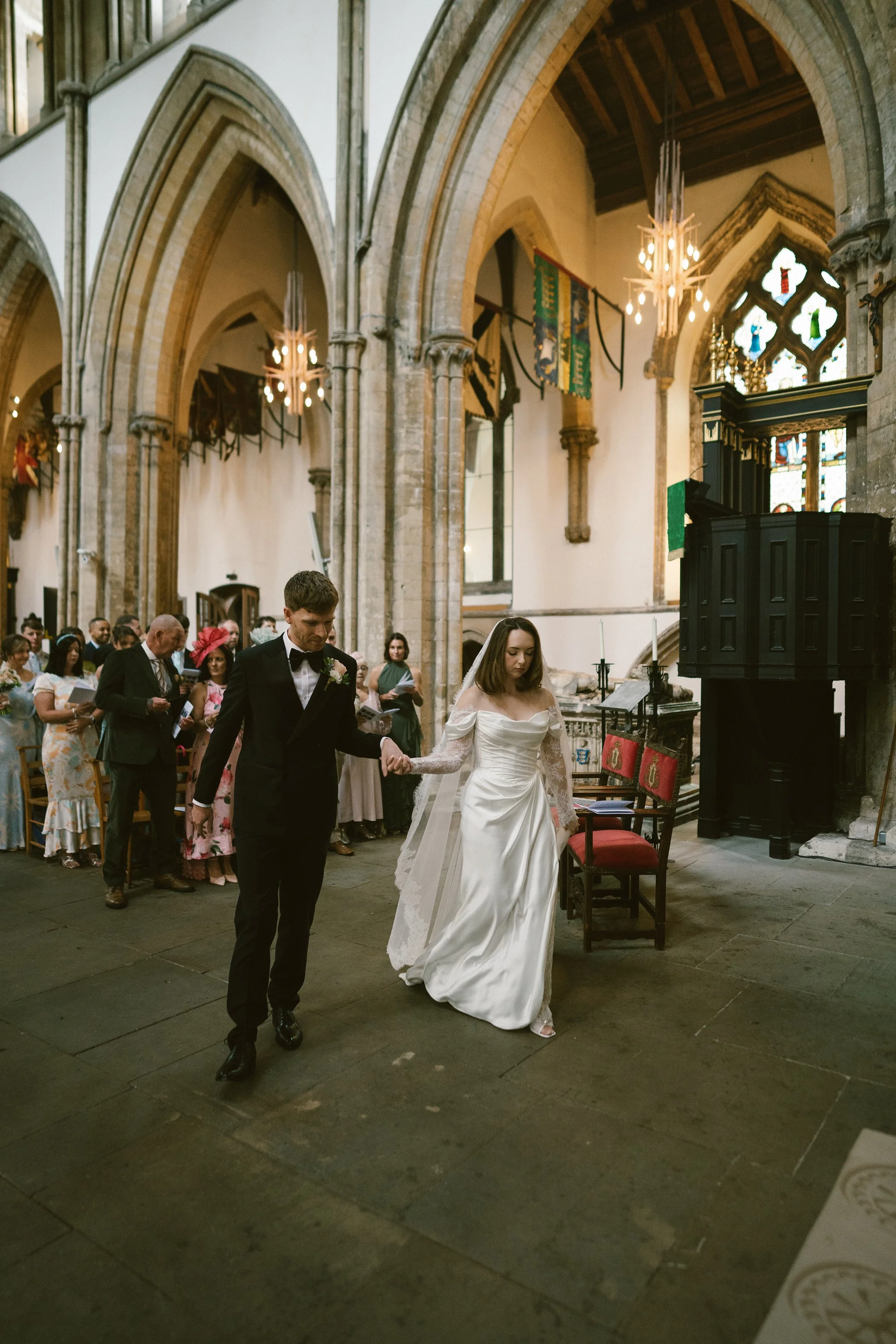 A bride and groom at a wedding ceremony inside a historic church. The bride is in a white gown, and the groom is in a black tuxedo. Guests are standing behind them, holding programs. The church has tall arched ceilings, stained glass windows, and dec