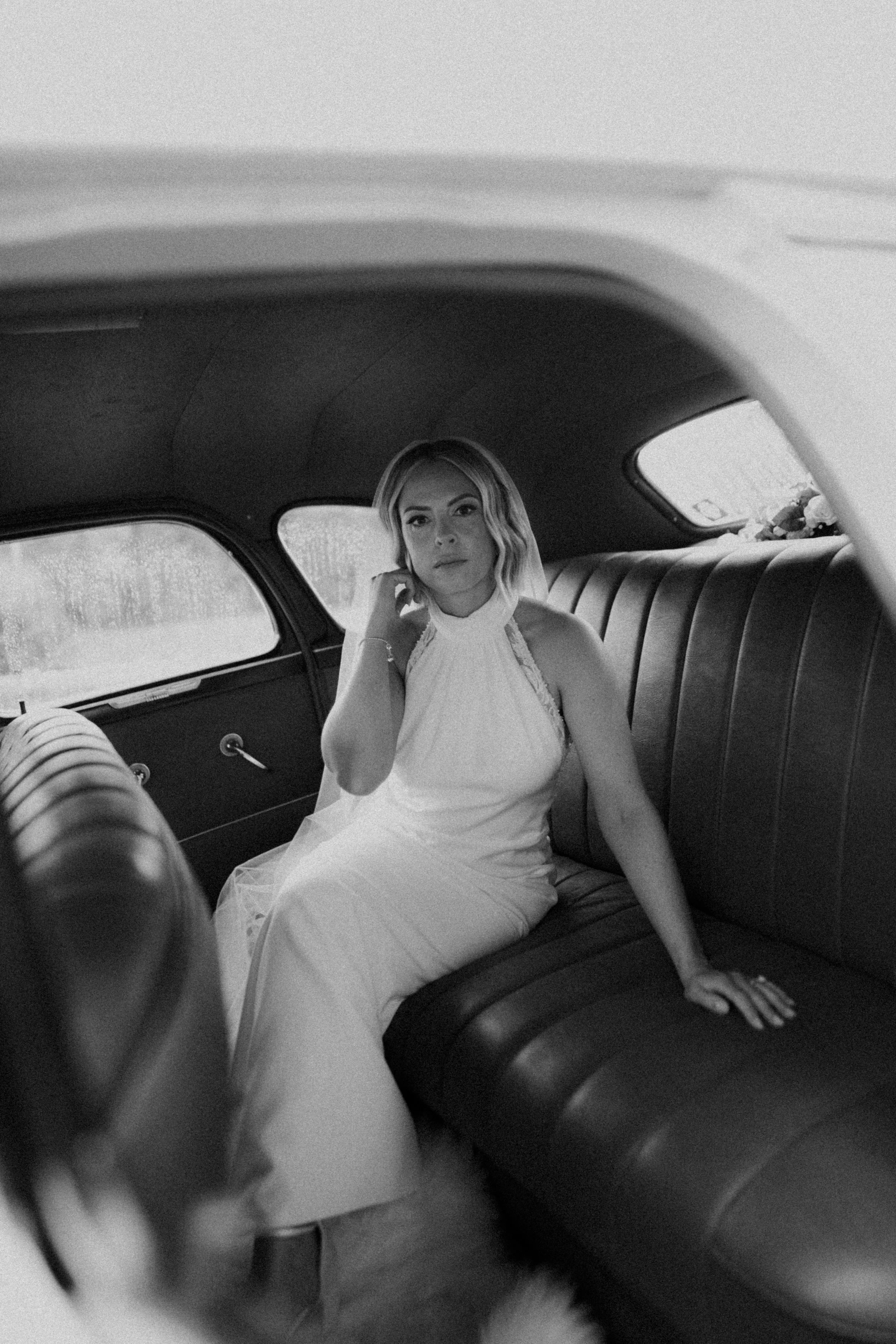 A woman in a white dress sitting in the back of a vintage car, looking at the camera with a contemplative expression.