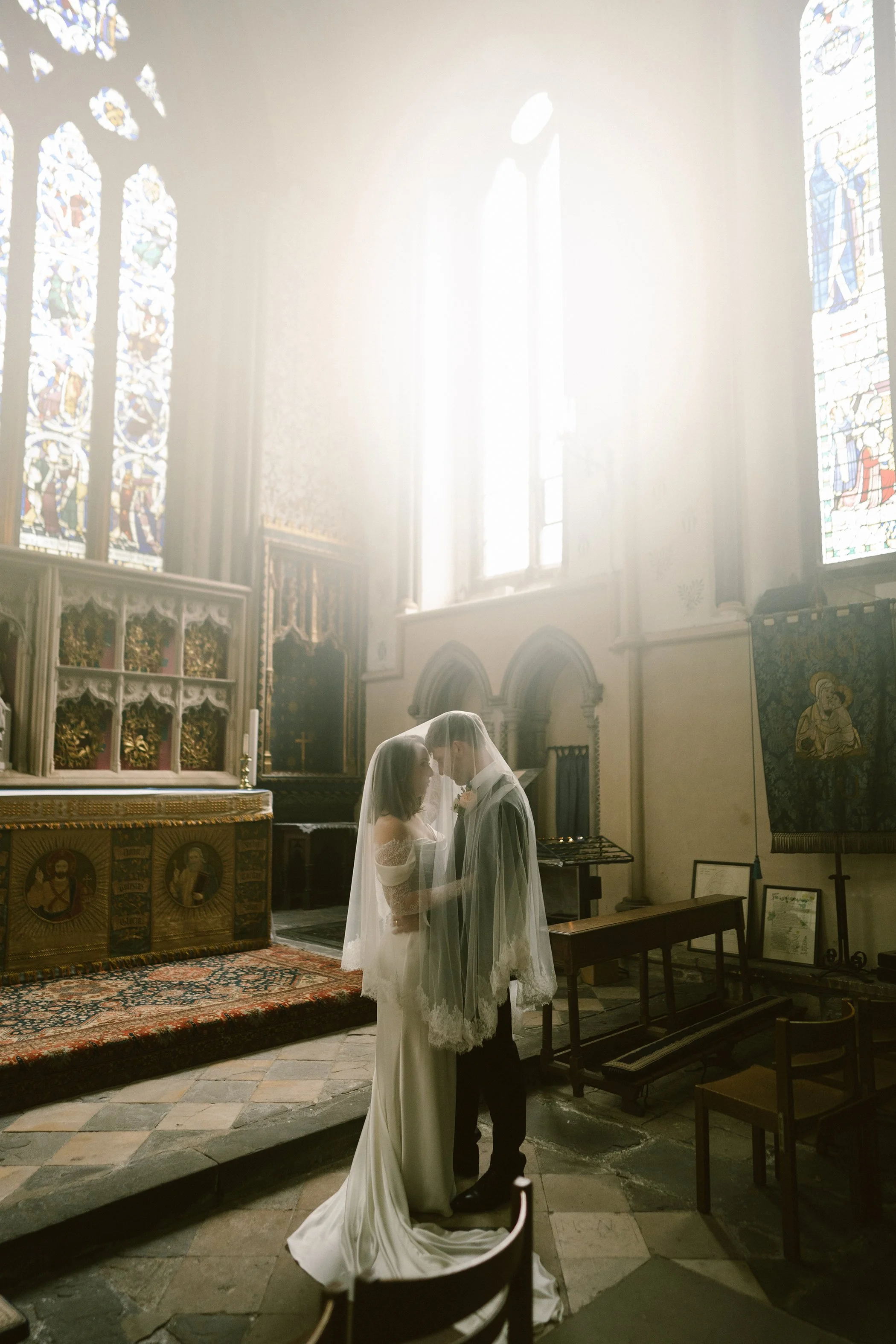 A bride and groom stand close together inside a church, holding hands and touching foreheads, during their wedding ceremony with stained glass windows in the background.