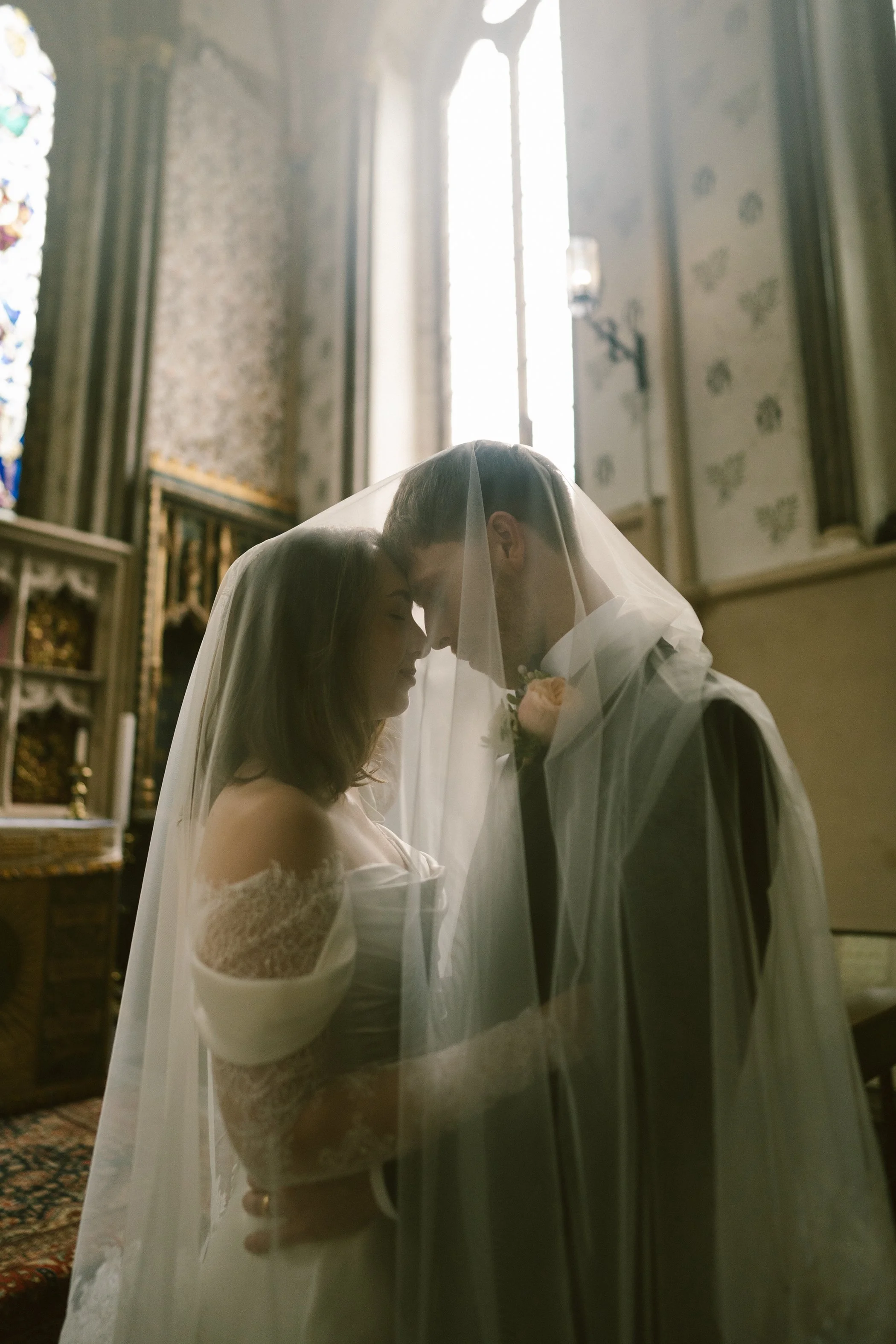 A bride and groom bow their heads together inside a church, with soft natural light coming through tall windows behind them. The bride is wearing a white lace dress and a veil, and the groom is dressed in a dark suit with a white shirt and tie. A bou