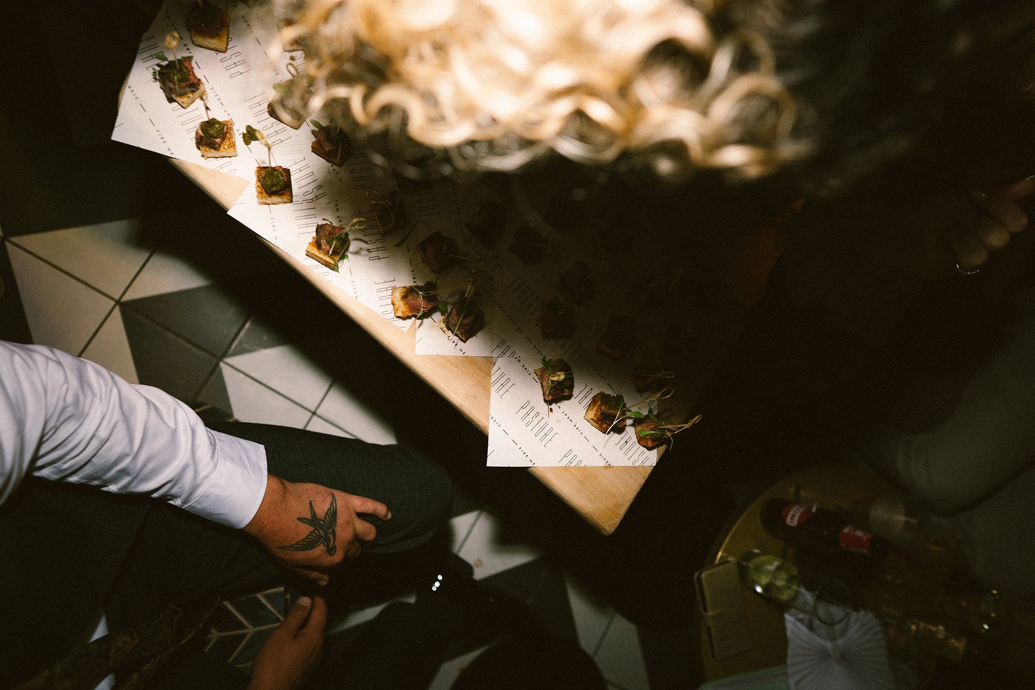 A close-up of a table with small appetizer bites topped with herbs, with a person’s arm and part of leg visible. The setting appears to be in a restaurant or bar with tiled flooring and a dark ambiance.