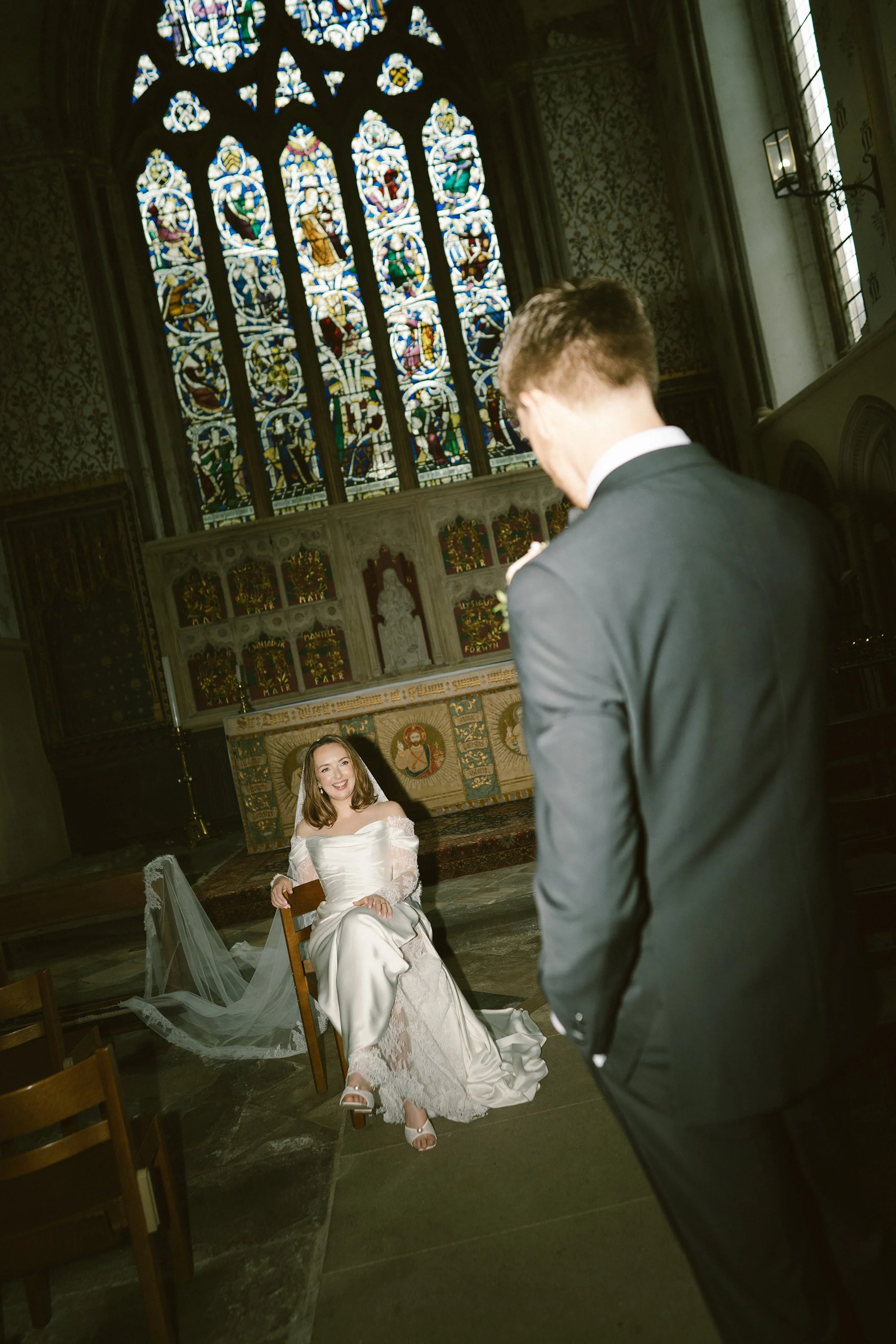A bride in a white wedding dress sitting in a church, smiling at a man in a black suit, inside a church with stained glass windows and religious artwork in the background.