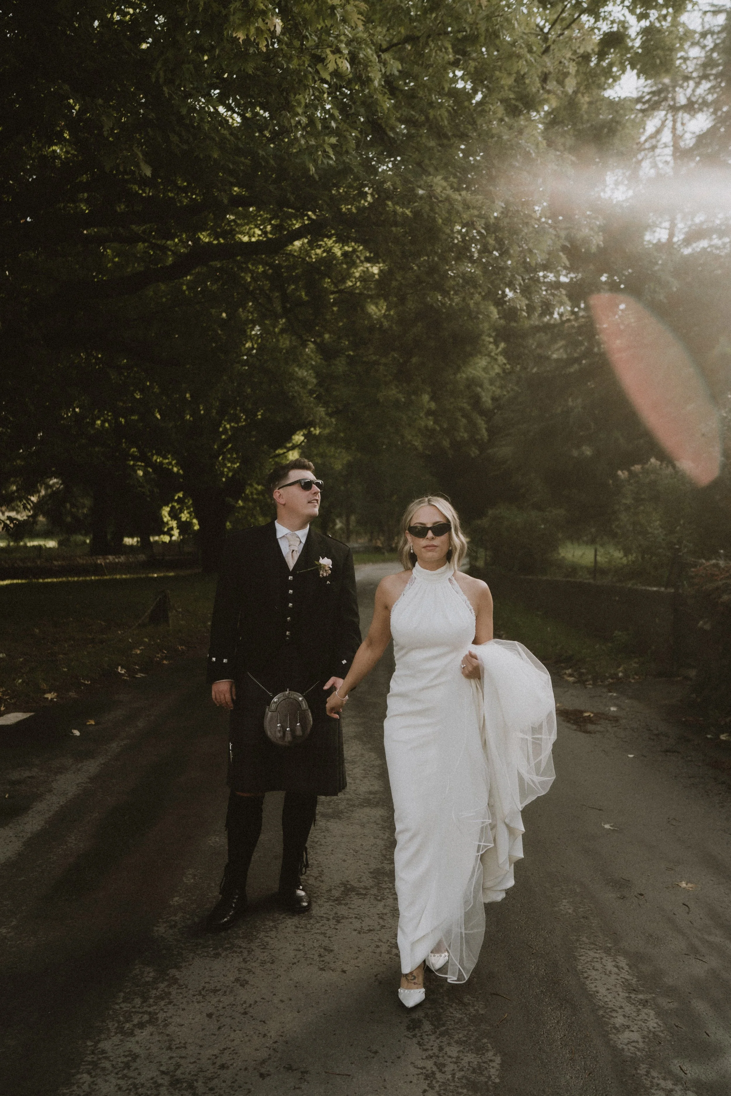 A couple, dressed in wedding attire and wearing sunglasses, walking hand in hand on a tree-lined road during daytime.