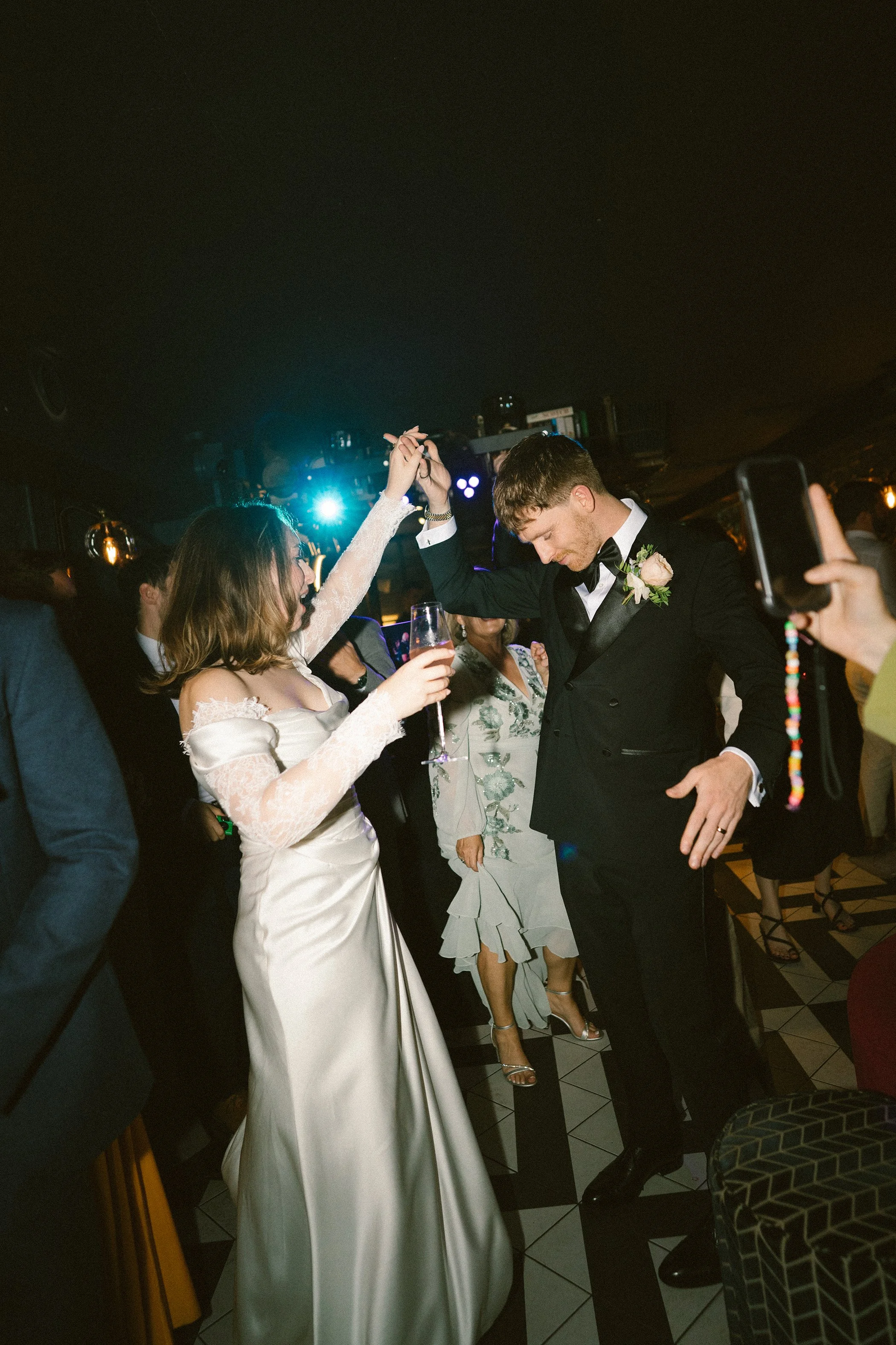 A groom and bride dancing at a wedding reception with guests watching. The bride is in a white wedding dress, and the groom in a black tuxedo. The setting has dim lighting, and there are people dressed formally, including women in dresses and men in 