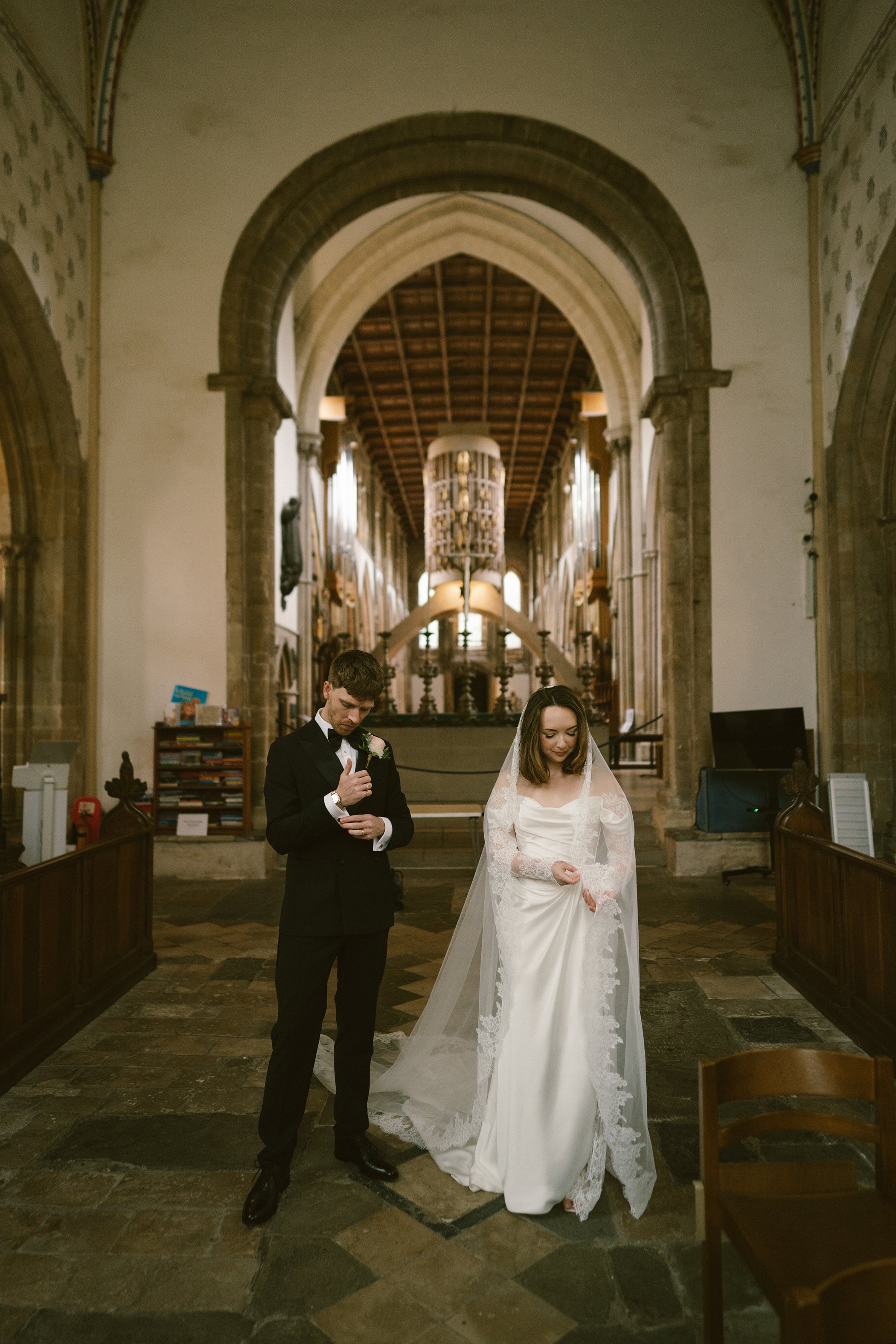 Bride and groom stand together in a church during a wedding ceremony, the groom in a black tuxedo with a boutonniere, the bride in a white wedding gown with lace details, both looking down, with an arched stone entrance and wooden ceiling in the back