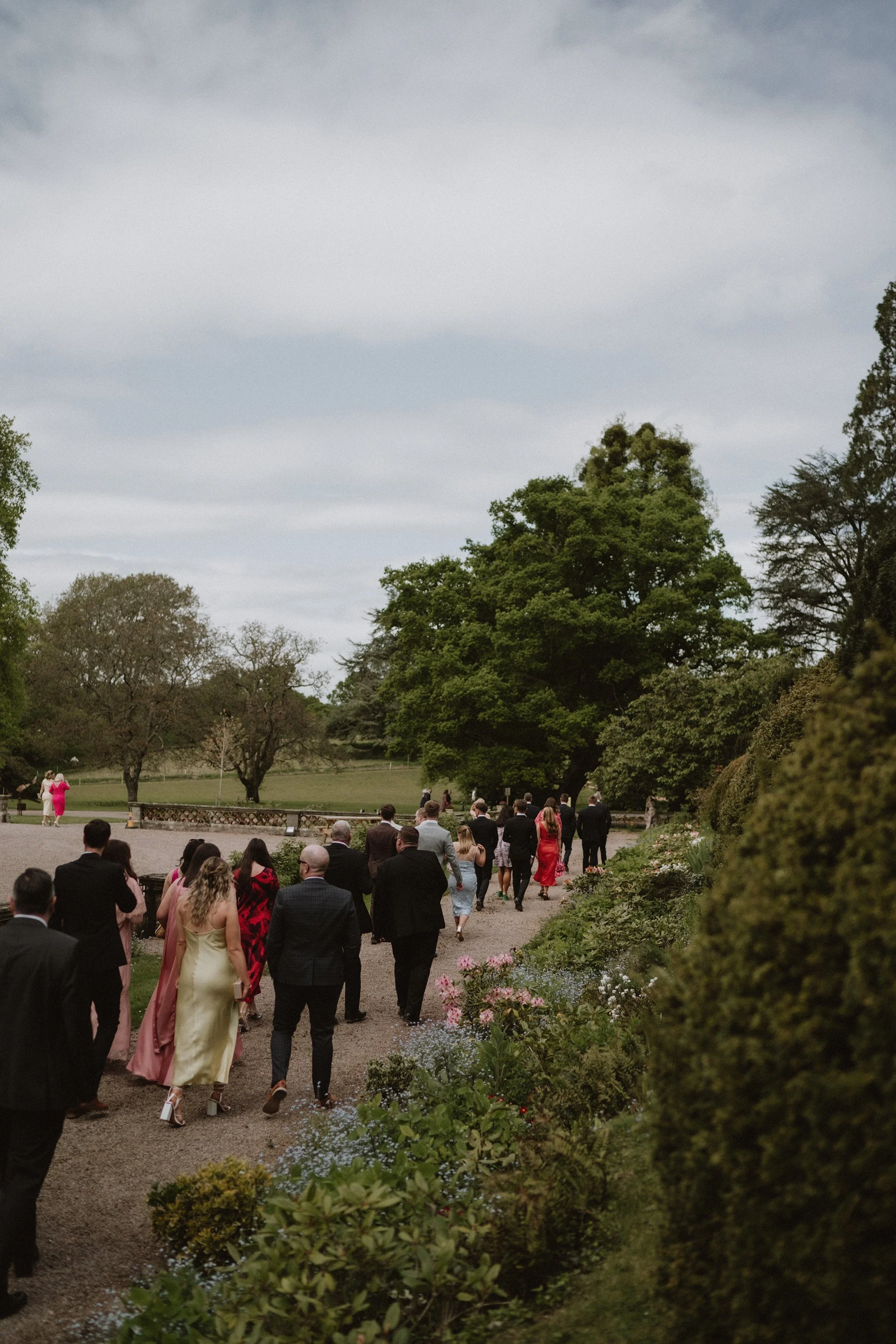People walking outdoors on a gravel path surrounded by greenery, with some dressed in formal attire, suggestive of a wedding or special event.