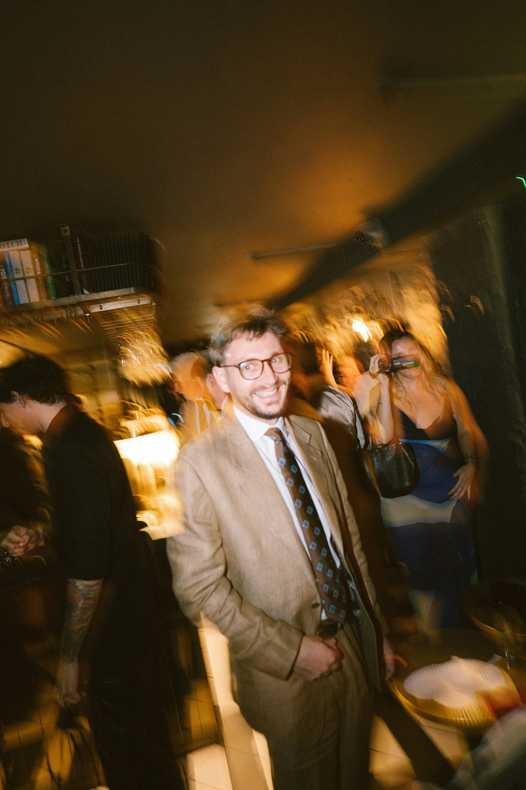 A man wearing glasses and a tan suit with a patterned tie smiling in a crowded indoor setting with warm lighting, others around him including a woman taking a photo.