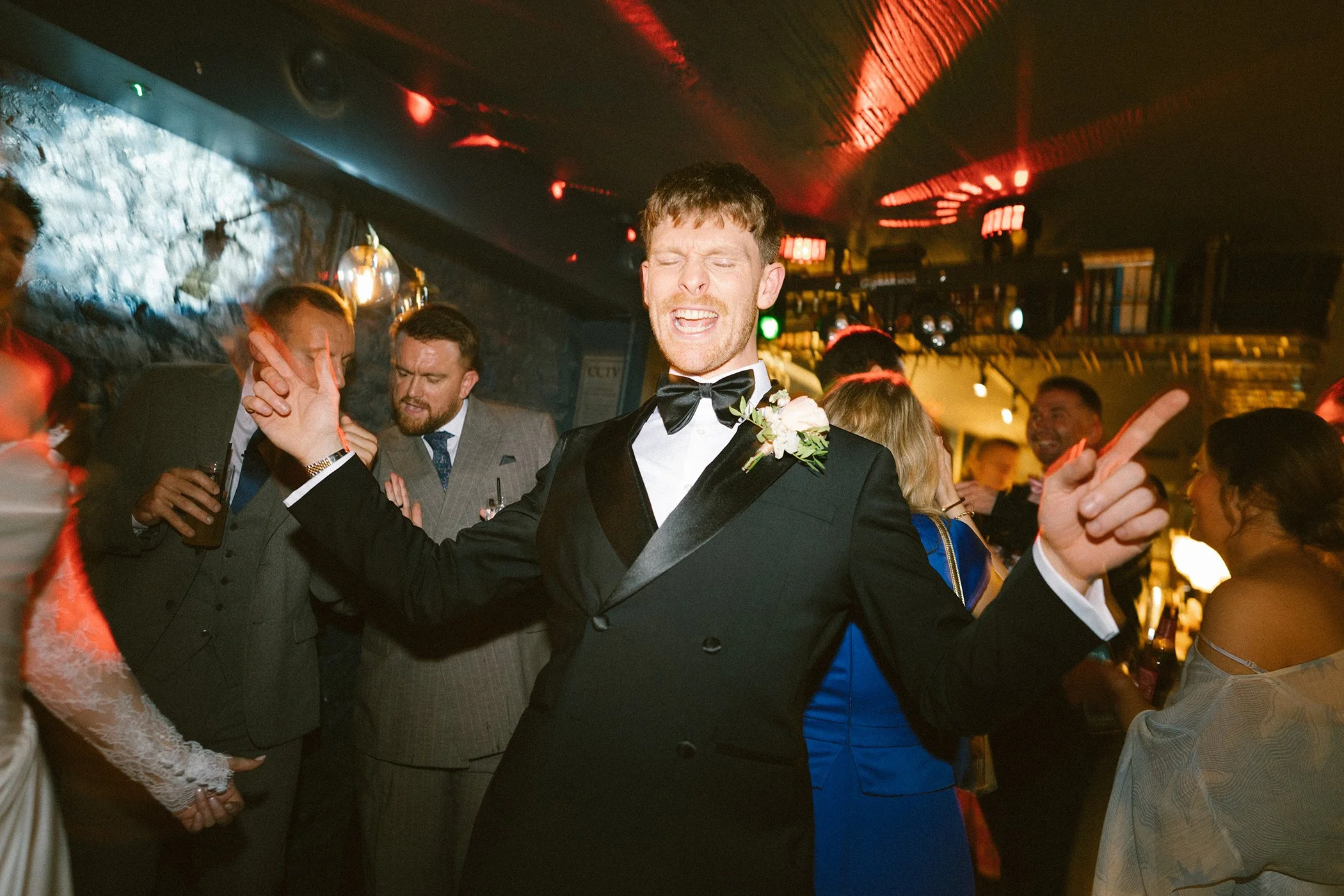 A groom in a tuxedo with a boutonniere dancing at a wedding reception with guests around him.