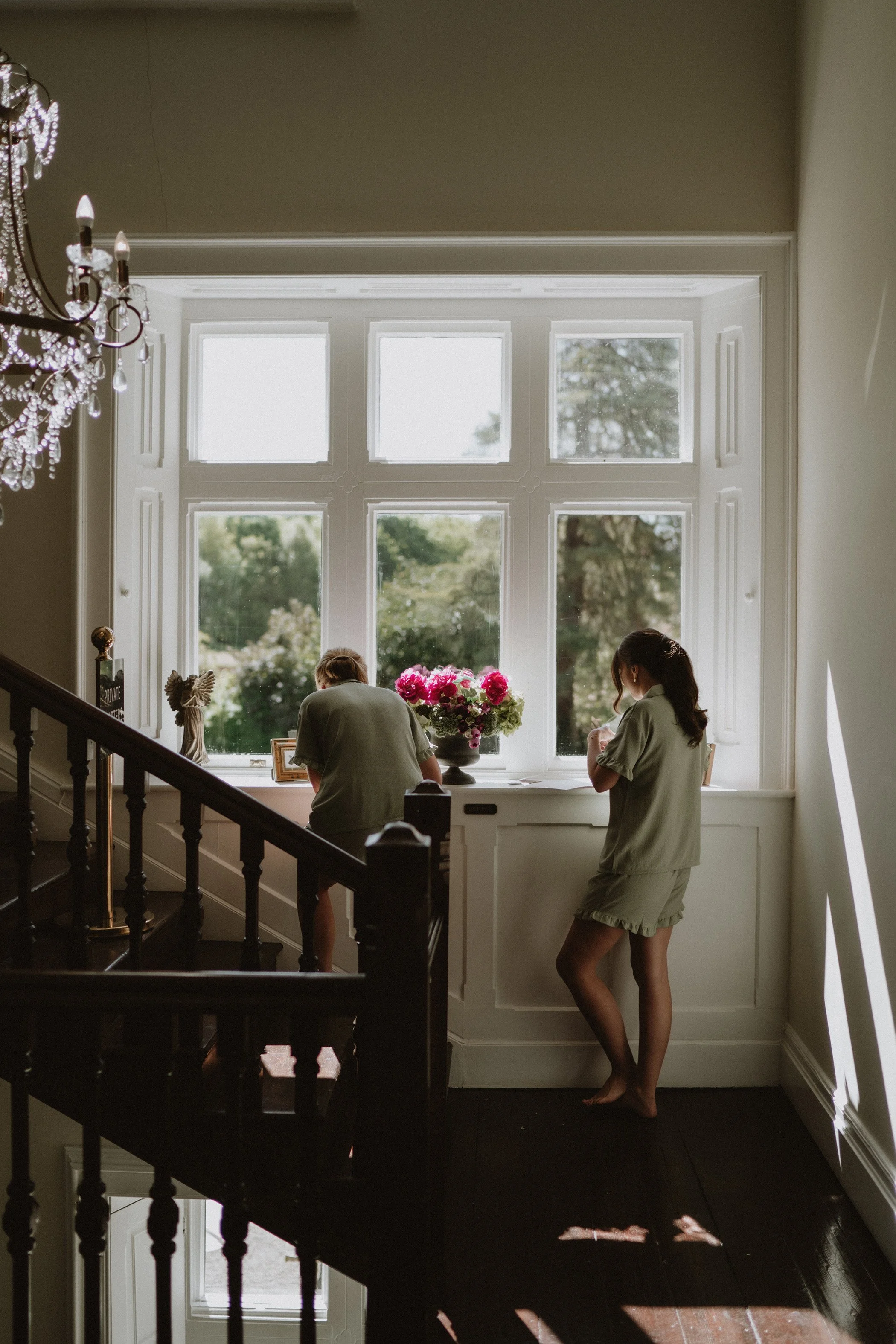 Two women in matching pajamas are standing by a large window with natural light, one looking down and the other turning slightly, with a vase of pink and white flowers between them inside a house.