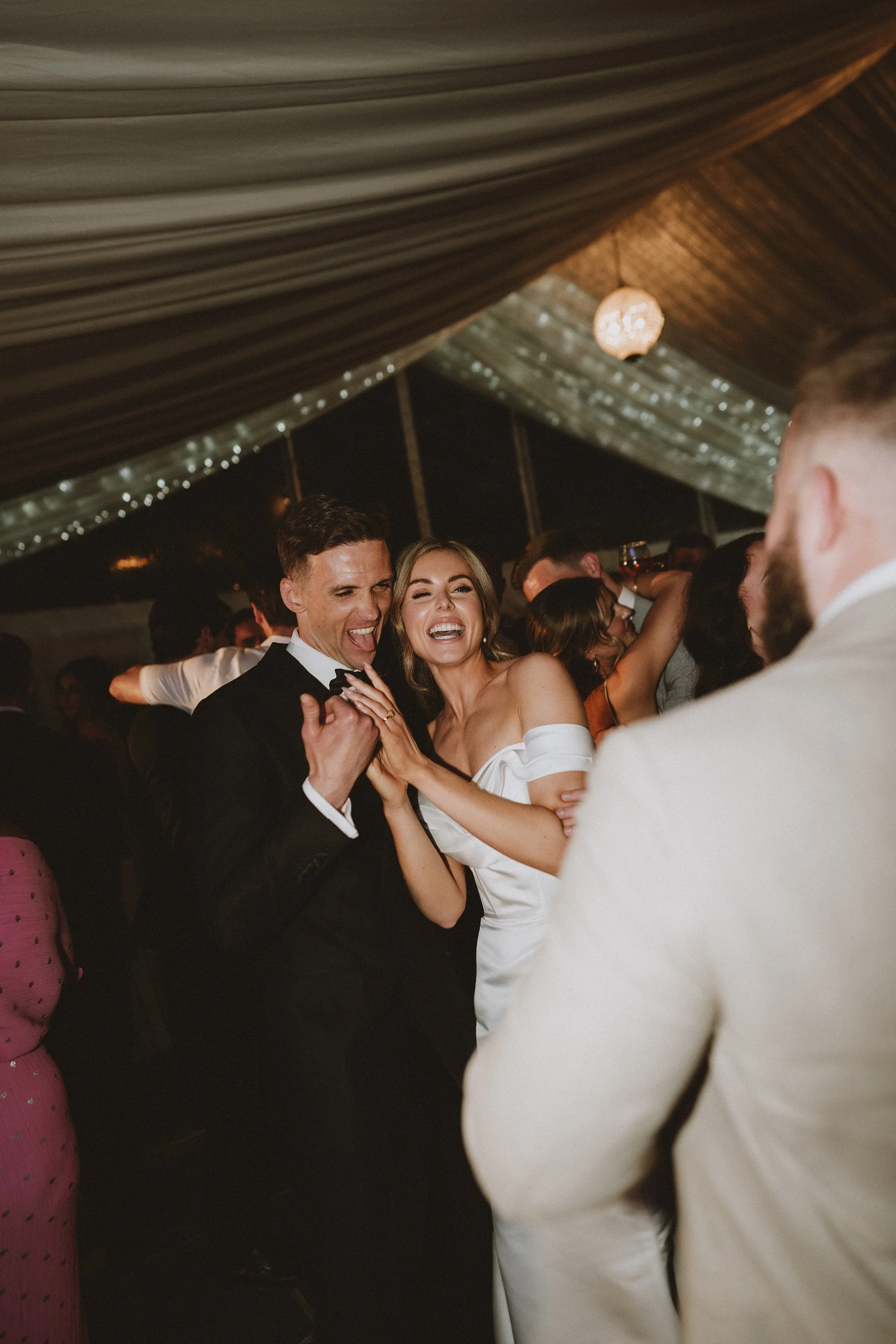 Happy bride and groom dancing and celebrating at their wedding reception with guests in the background under decorated tent.