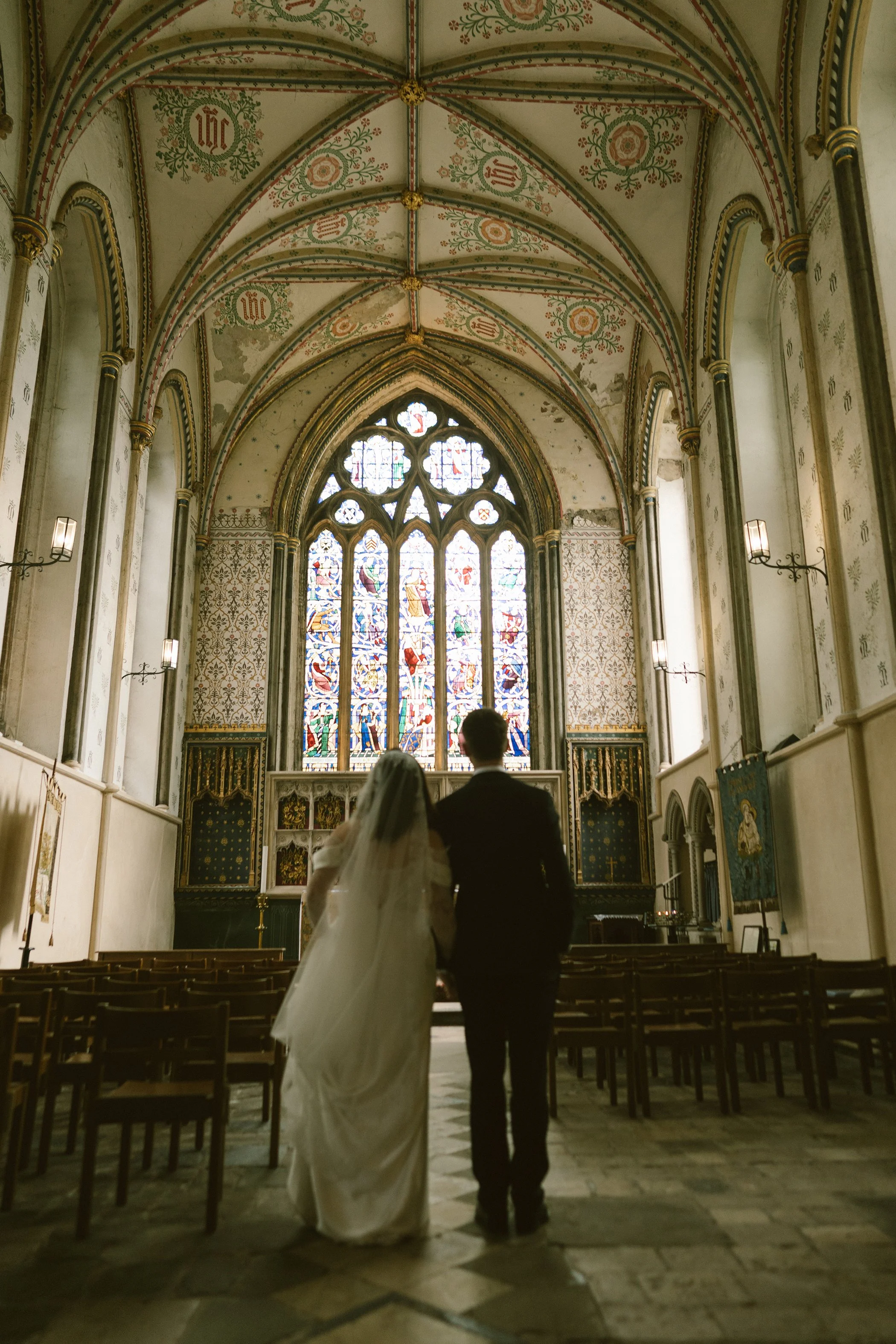 A bride and groom walking down the aisle in a church with stained glass windows, ornate ceiling, and wooden pews.
