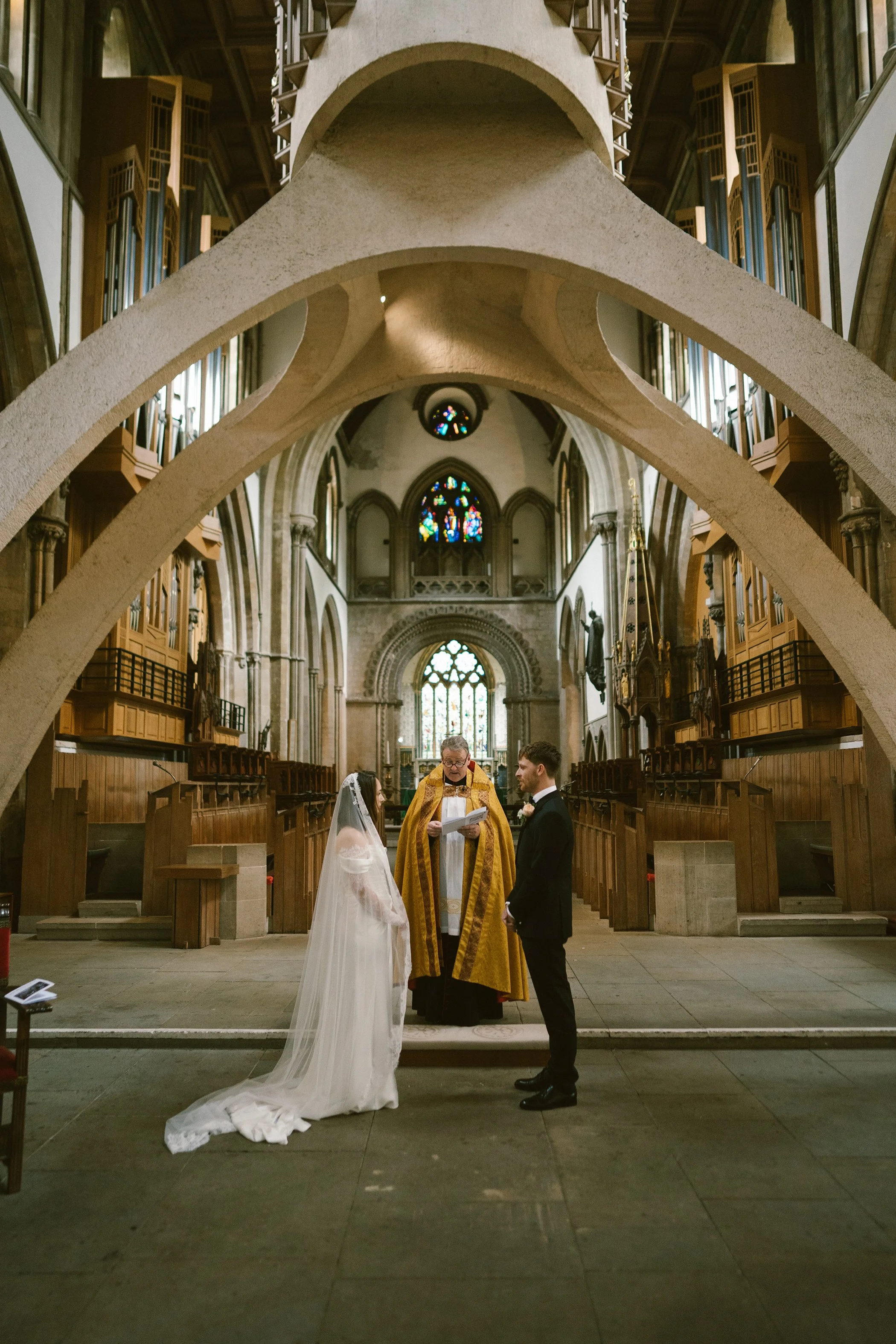 A wedding ceremony taking place inside a church, with a bride and groom standing before a priest. The church features large stained glass windows and gothic architecture.