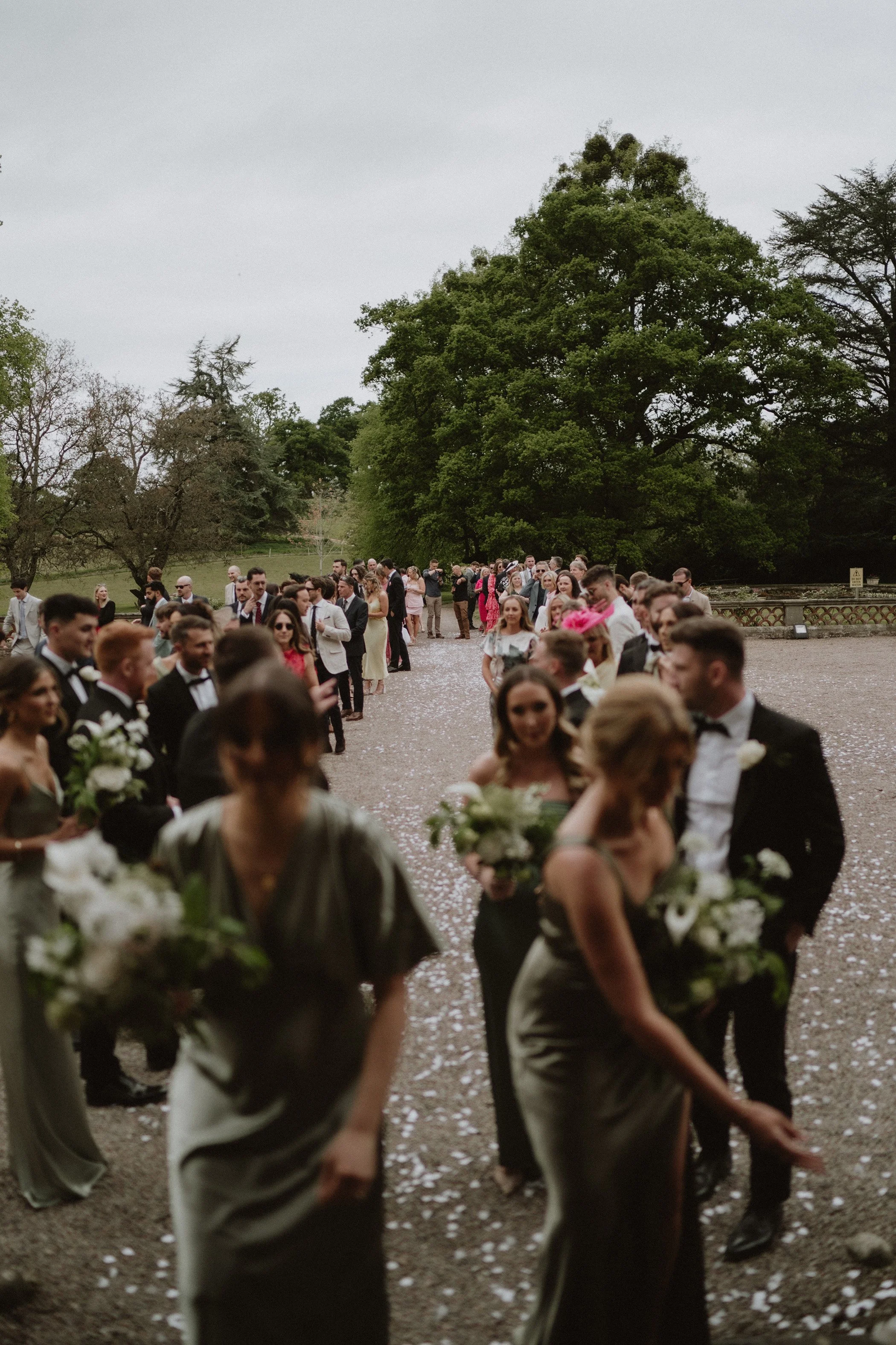 Groups of people dressed in formal attire, including tuxedos and dresses, participating in an outdoor wedding ceremony in a lush green park surrounded by trees and falling flower petals.