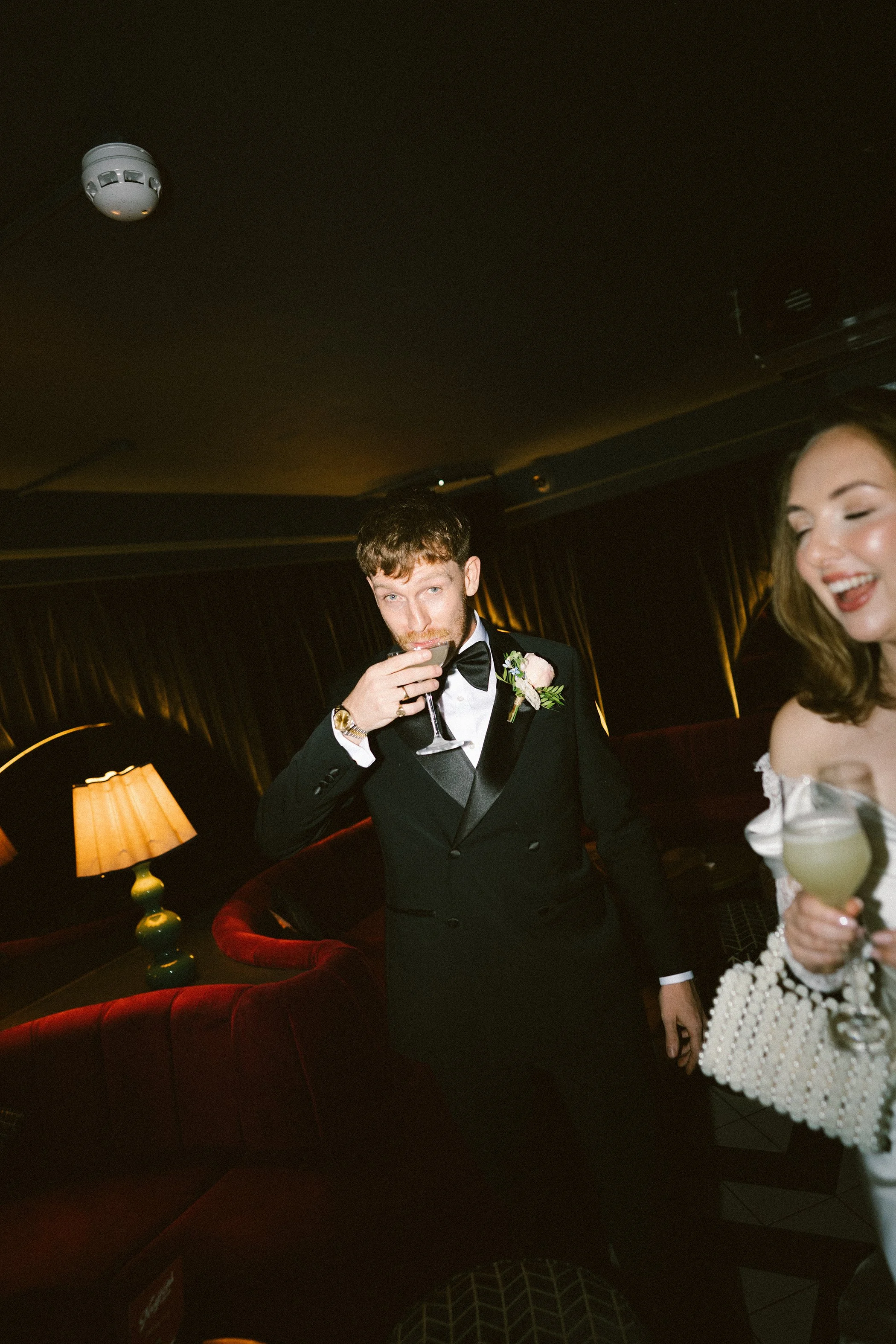 A man in a tuxedo drinking from a glass and a woman in a white dress smiling at an evening event.
