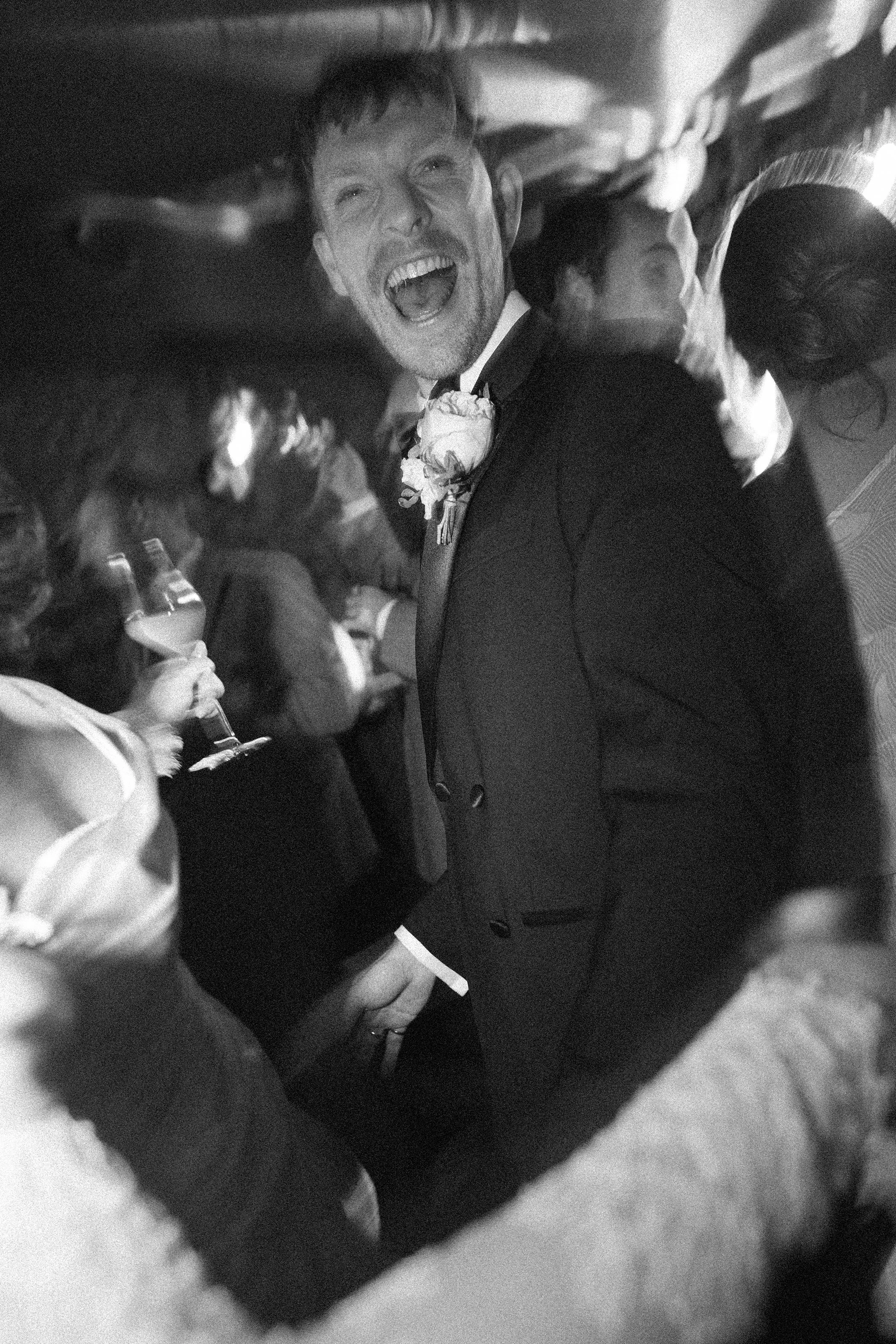 A black and white photo of a man in a tuxedo laughing joyfully at a wedding or celebration event, surrounded by other people.