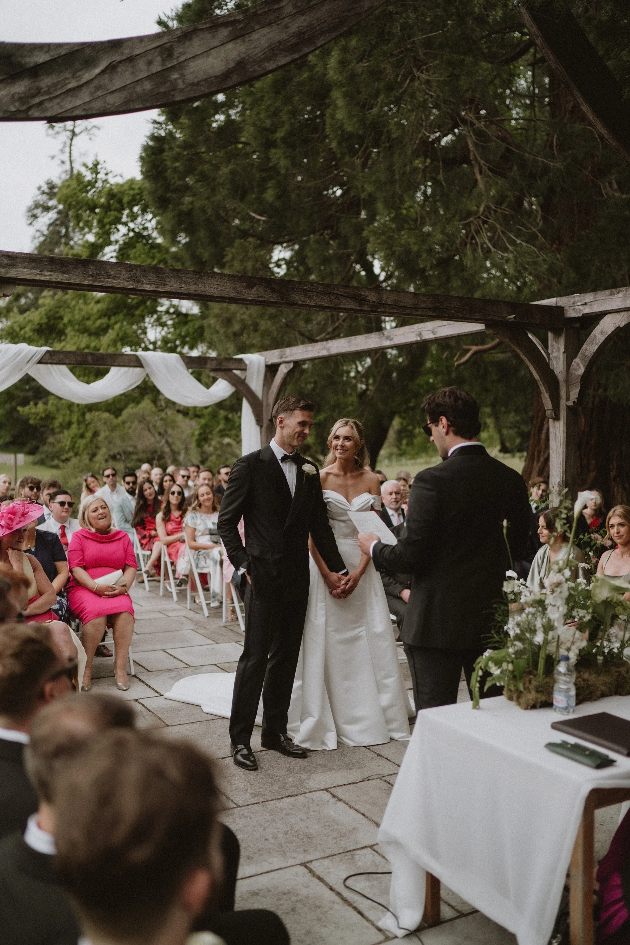 A wedding ceremony outdoors with a bride and groom holding hands, standing before an officiant, surrounded by seated guests under a wooden arbor with white drapery and greenery.
