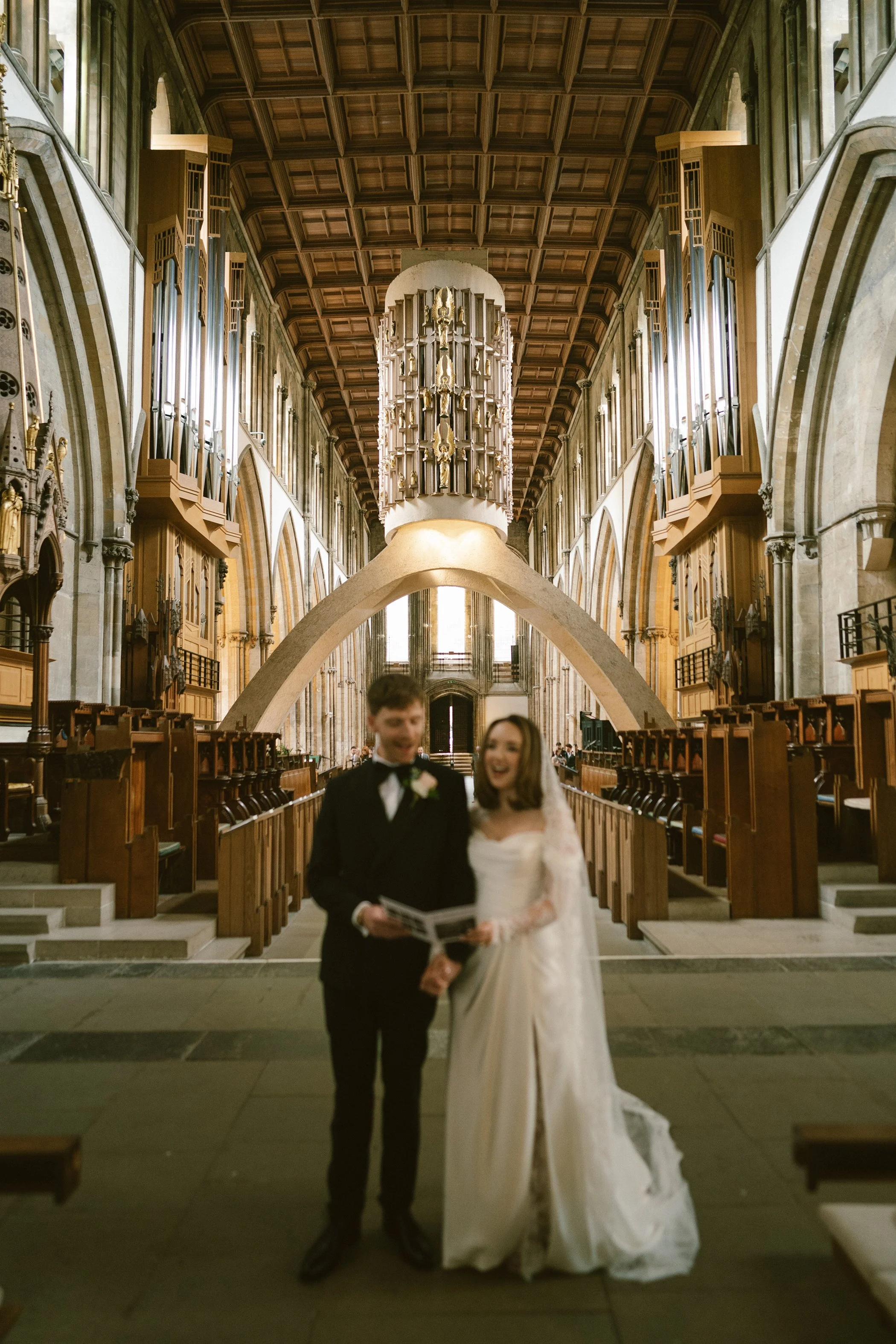A bride and groom stand inside a church with wooden pews and high arched ceilings. The bride wears a white dress with lace long sleeves, and the groom wears a black tuxedo with a white shirt and black bow tie. They are smiling and holding a booklet o