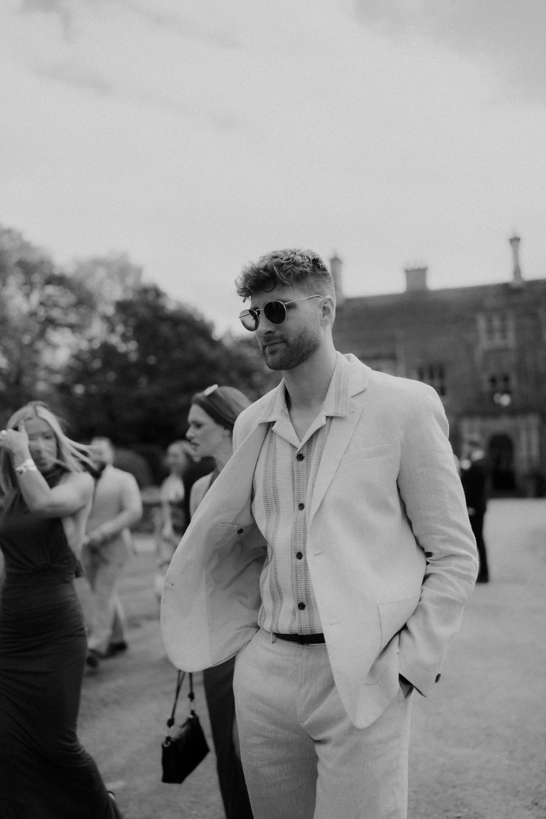 A man wearing sunglasses, a light-colored suit, and a patterned shirt, standing with his hands in his pockets, is in an outdoor setting with people in the background.