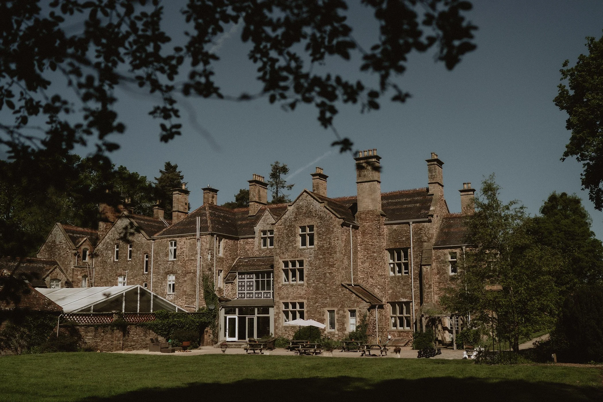 Large historic brick mansion with multiple chimneys, surrounded by trees and a lawn, under a clear blue sky.