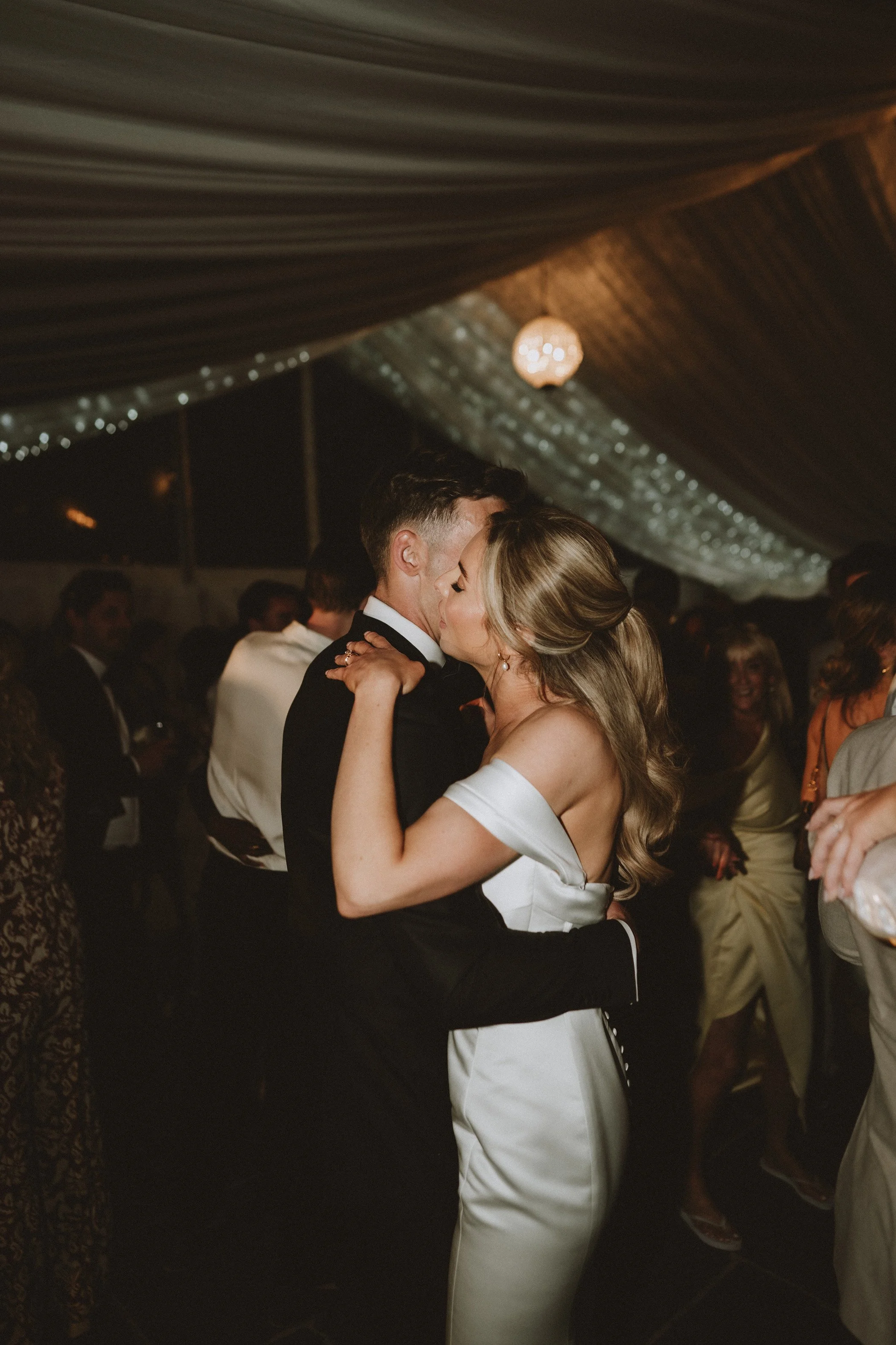 A couple dancing closely at a wedding reception, surrounded by guests in a dimly lit venue with hanging lights and draped ceiling.