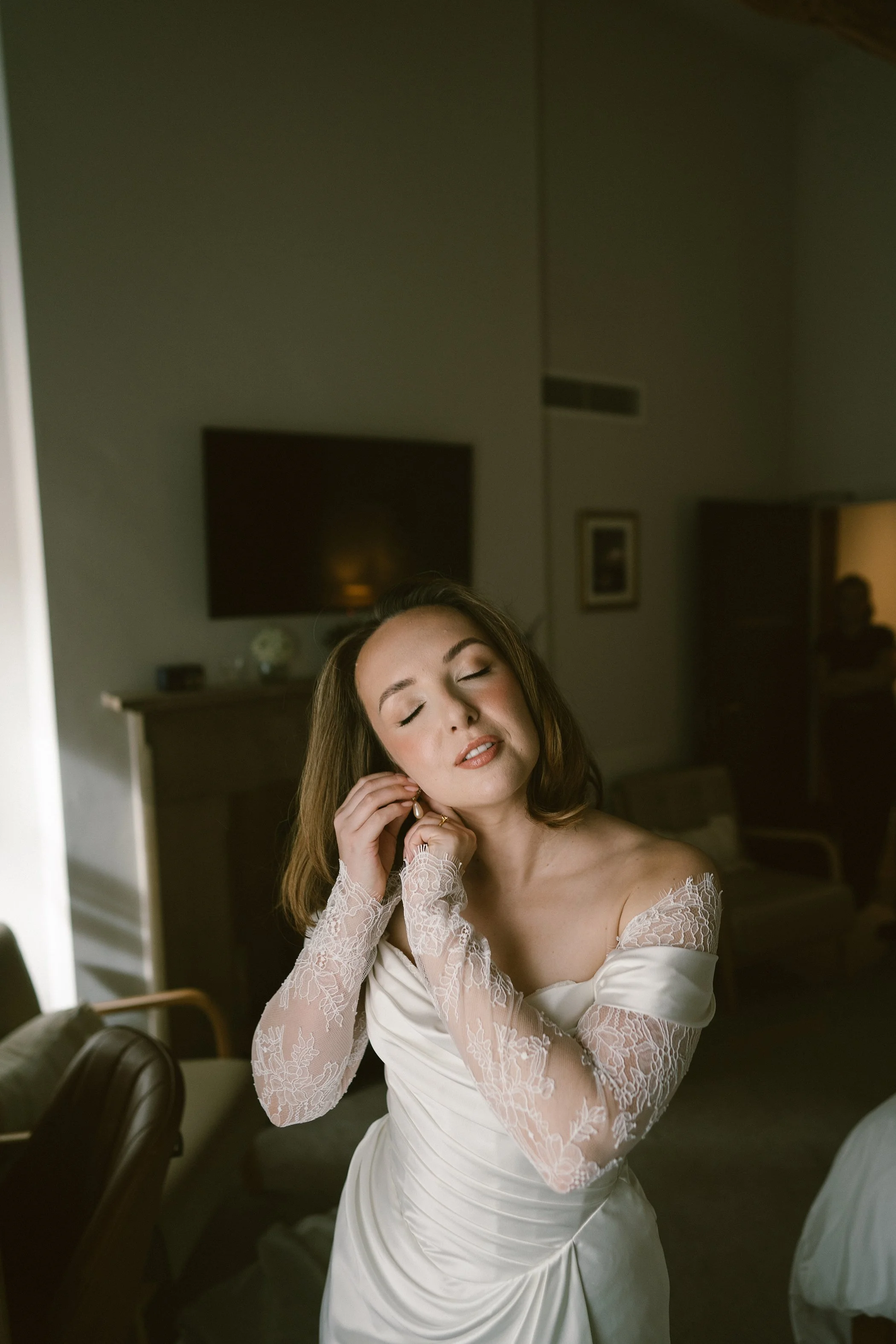 A woman with closed eyes in a wedding dress, adjusting her earring with lace long sleeves in a dimly lit room.