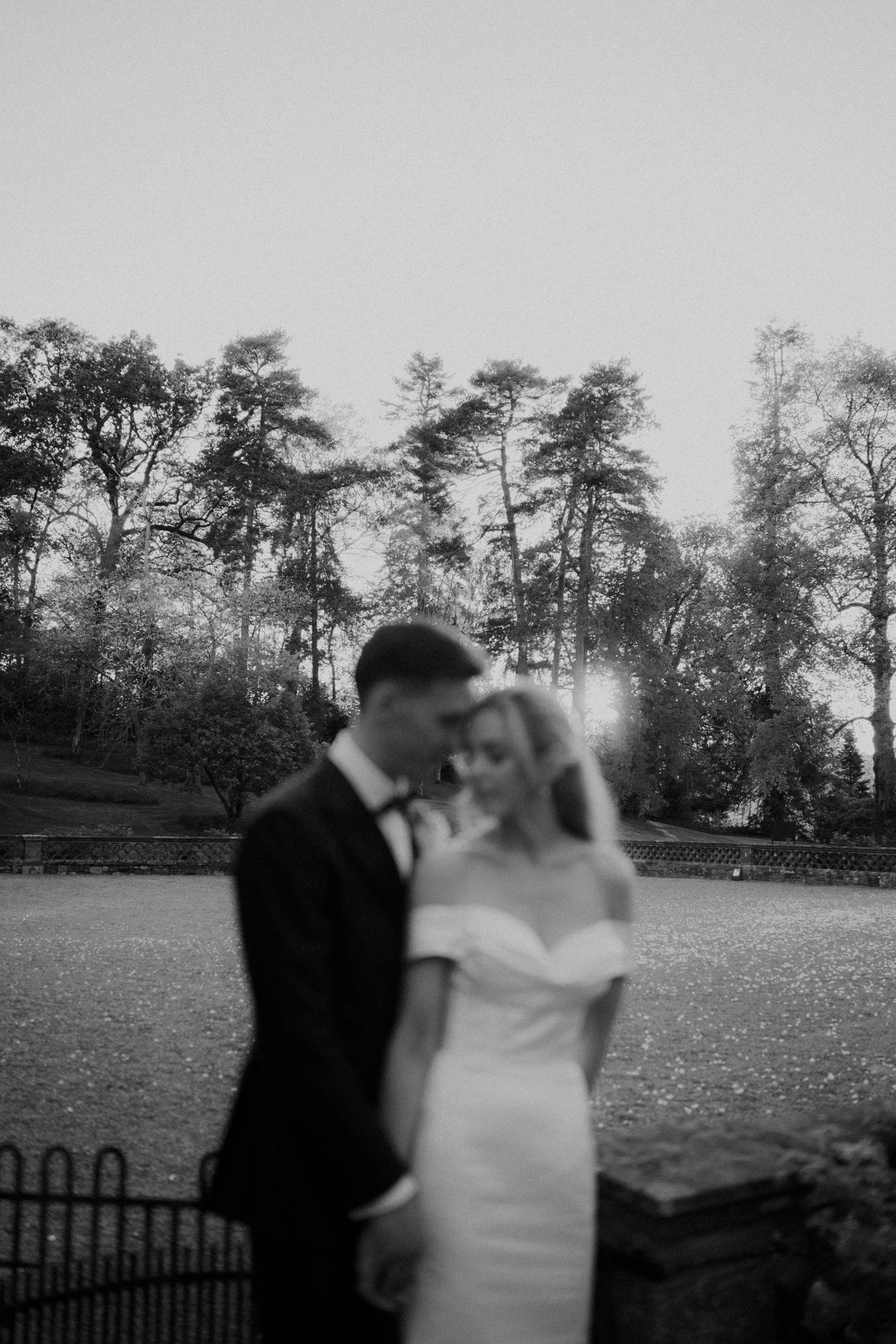 Blurred black-and-white photo of a wedding couple standing outdoors with trees in the background, the groom in a tuxedo and the bride in an off-shoulder wedding dress, holding hands.