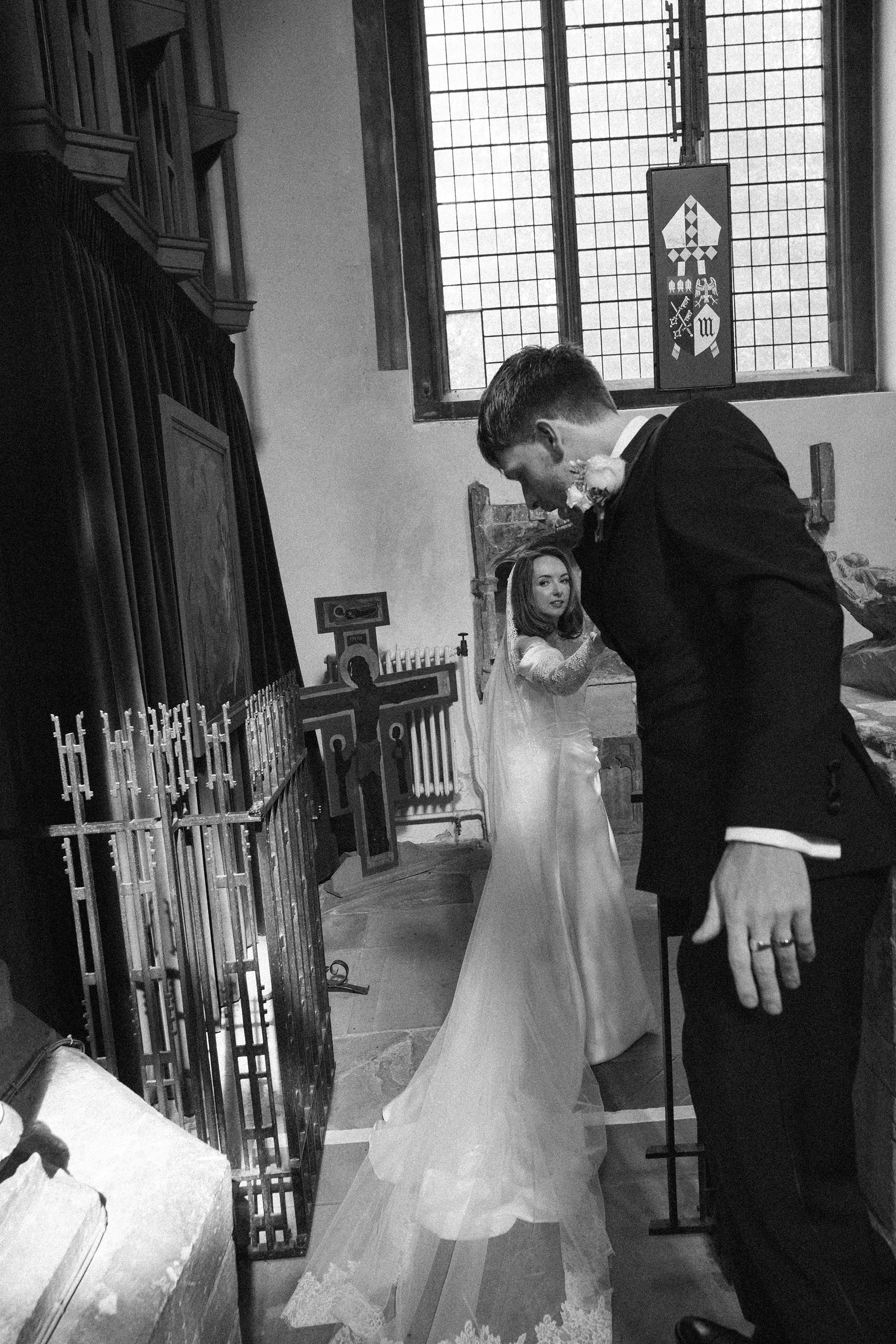 A black and white photo of a couple during a wedding ceremony inside a church. The groom is bowing his head and holding the bride's hand, who is looking at him. The bride is wearing a white wedding dress with lace, and the groom is in a dark suit. Th