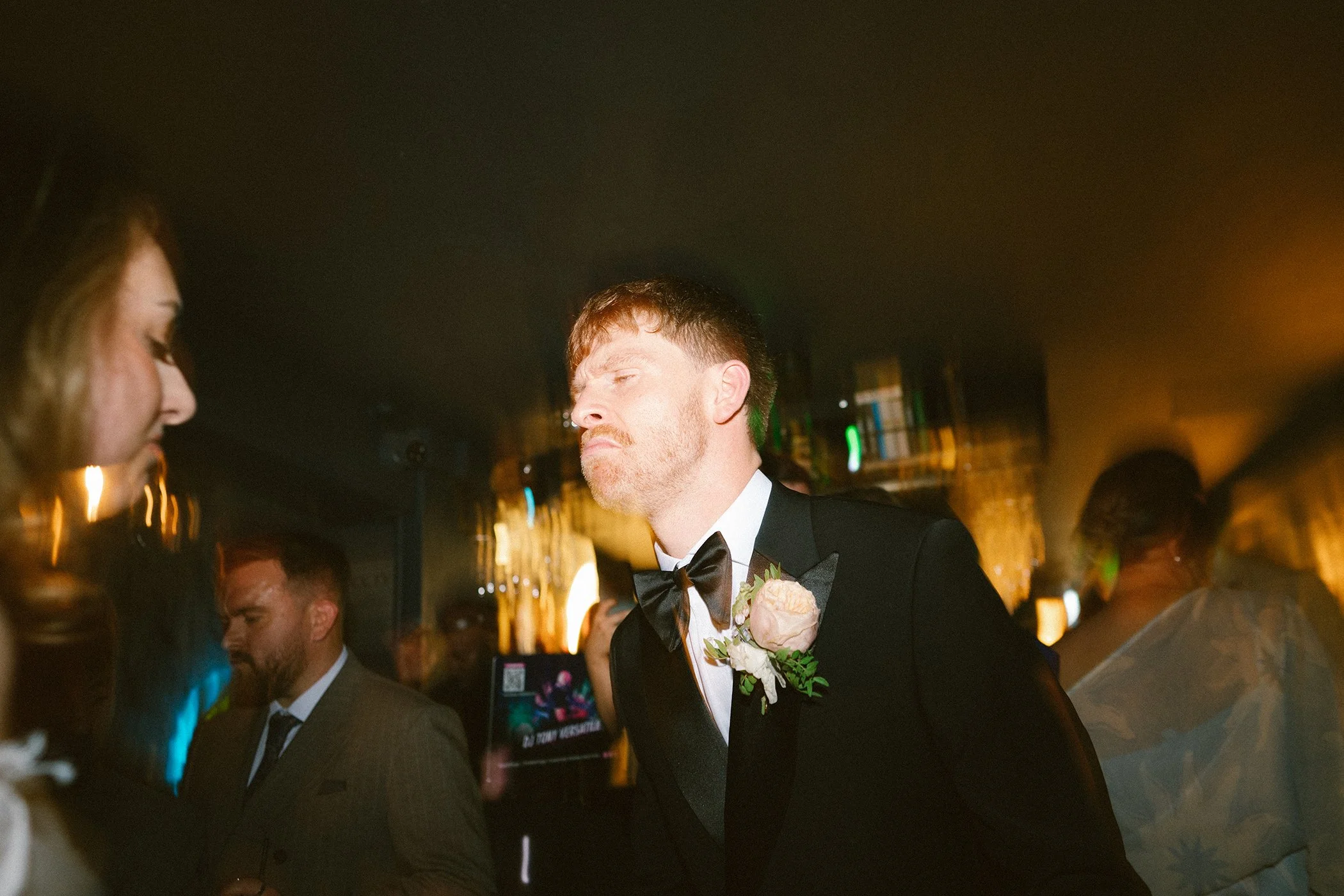 A man in a tuxedo with a boutonniere, possibly at a wedding, standing among a crowd at a formal event with a woman on the left side of the image, blurred background with warm lighting.