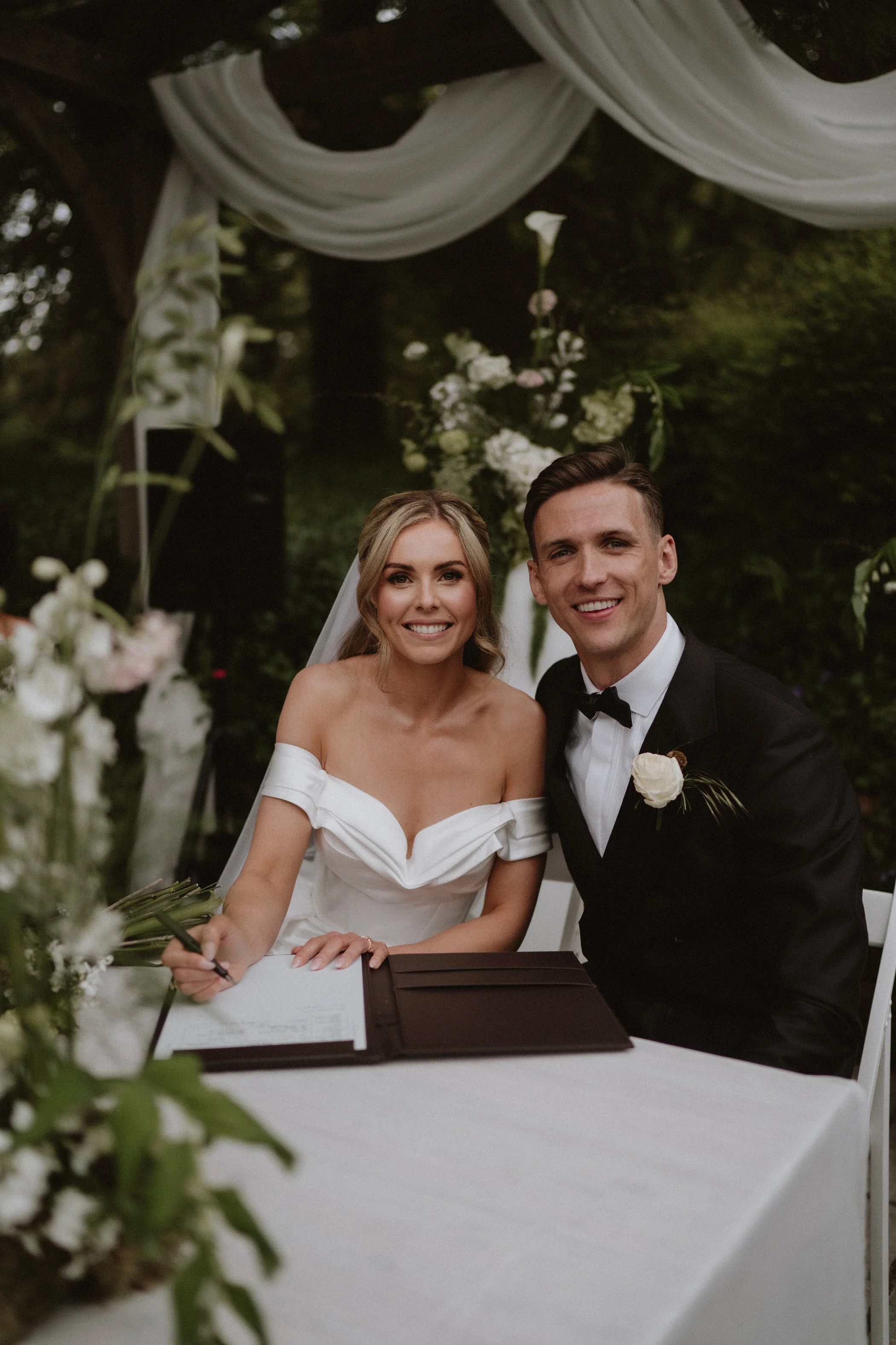 A smiling bride and groom sitting at a table outdoors, signing a marriage certificate, surrounded by floral decorations and greenery.