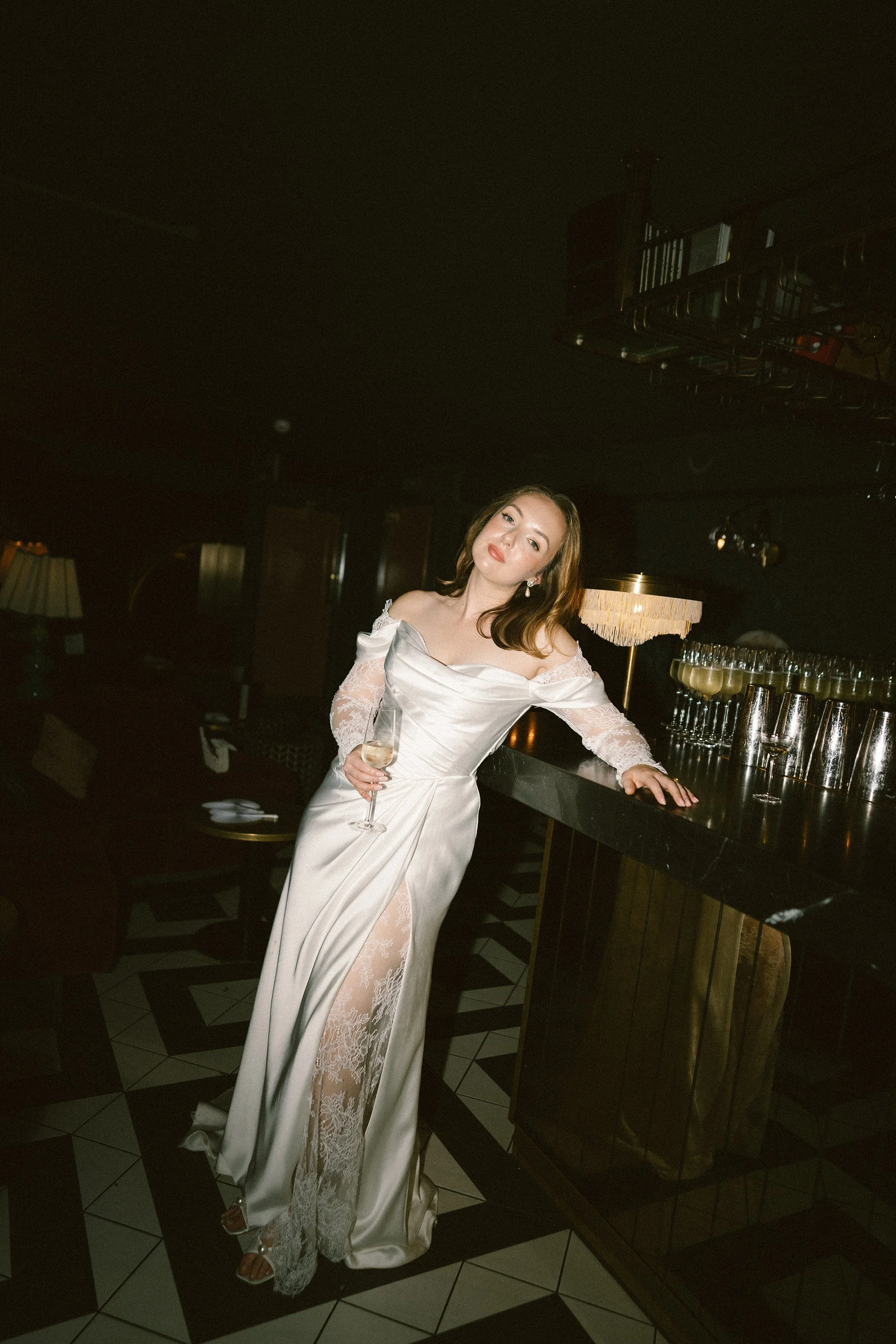 Woman in an elegant off-shoulder satin gown with lace details, holding a glass of champagne, standing at a bar in a dimly lit elegant venue.