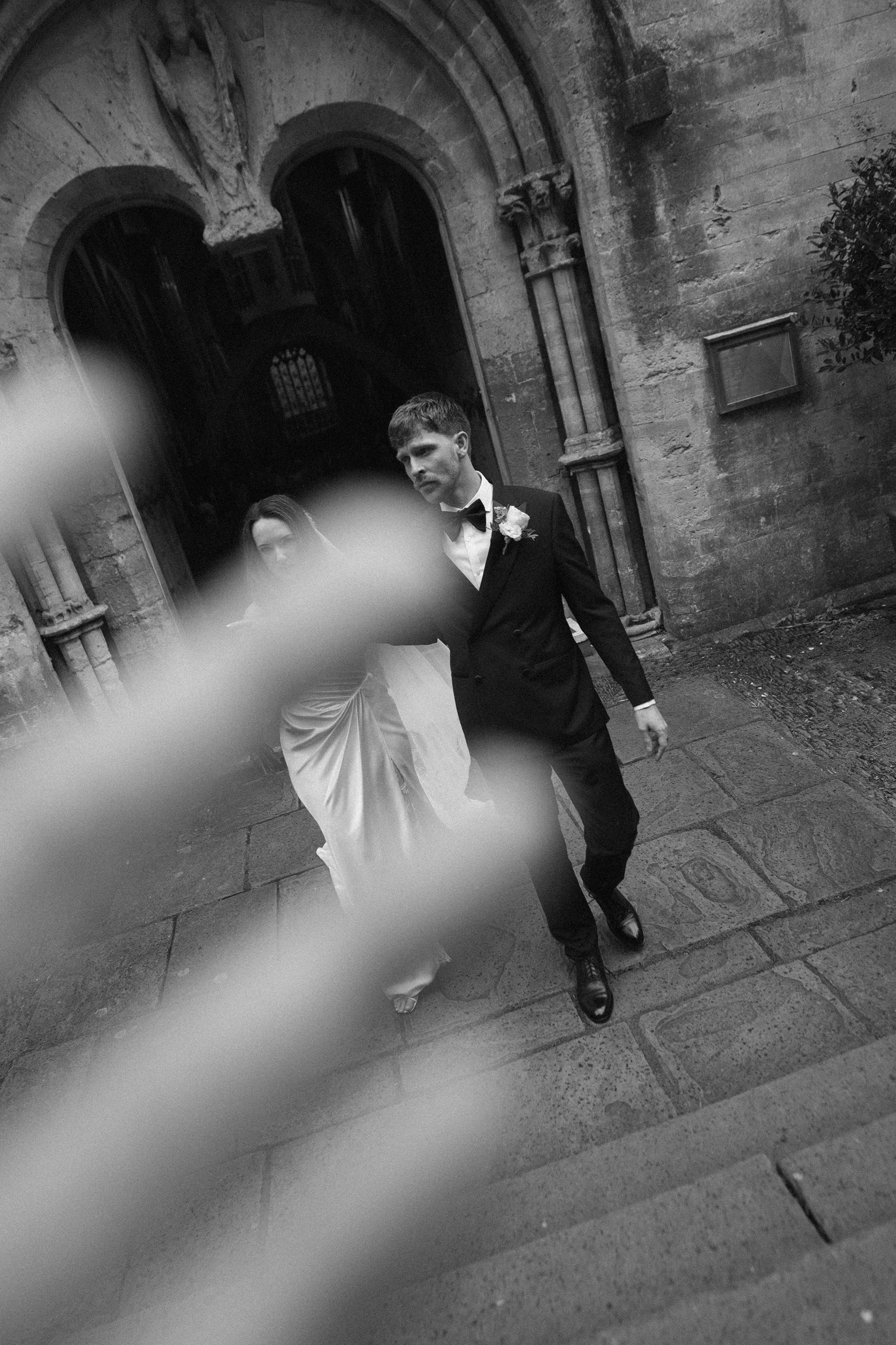 A black and white photo of a couple dressed in formal wedding attire, walking together outside a stone building with arched doorways and decorative architectural features. The man is wearing a tuxedo with a bow tie, and the woman is wearing a wedding