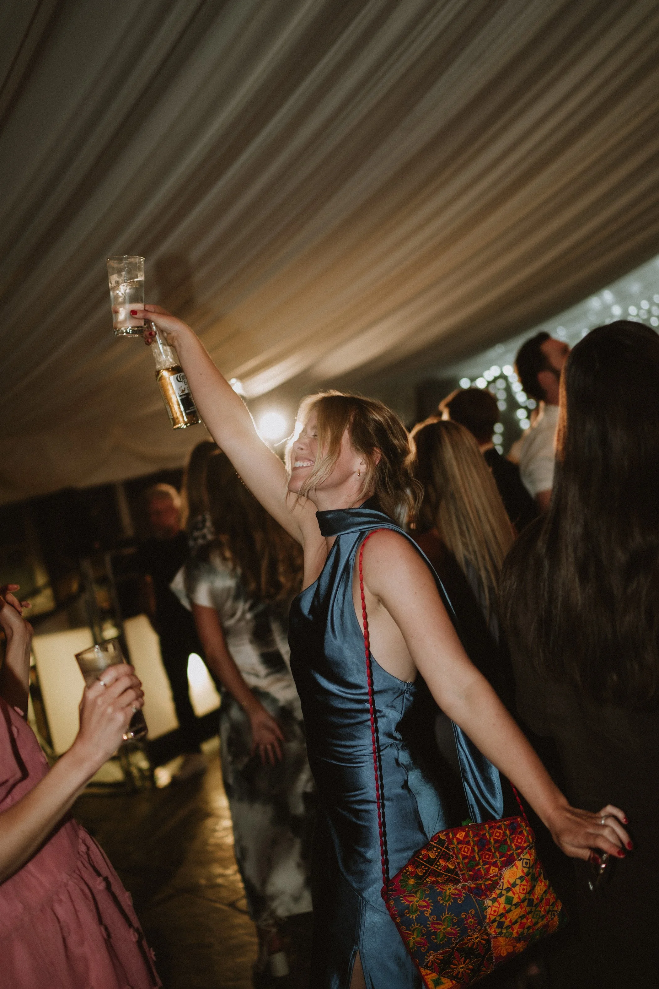A woman in a shiny blue dress at a party raising a glass in a toast, surrounded by other people dancing and celebrating under a decorated ceiling.