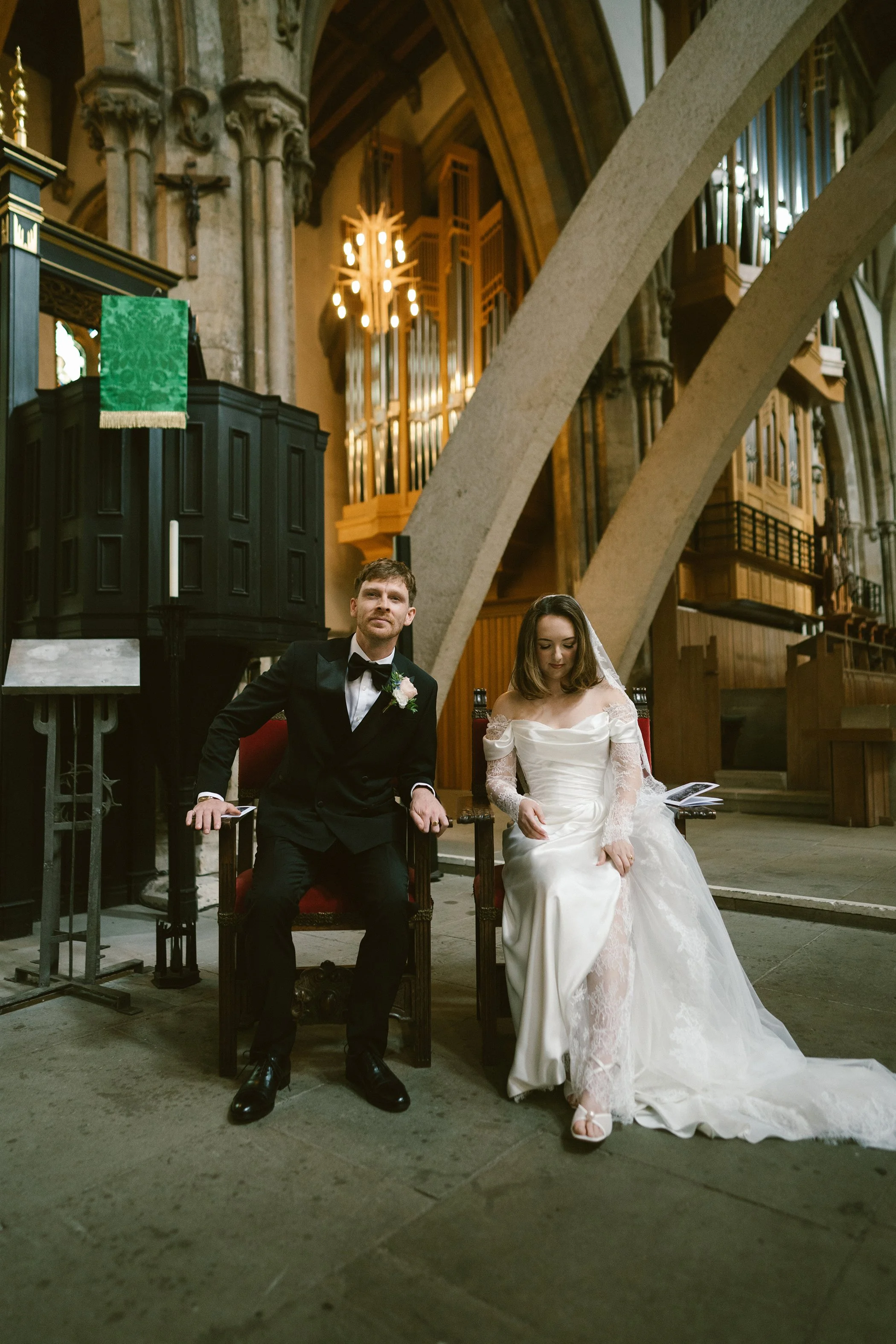 A bride and groom seated in a church during their wedding ceremony, with the groom in a black tuxedo and the bride in a white wedding gown with lace sleeves. The church features altar railings, an organ, and high stone arches.