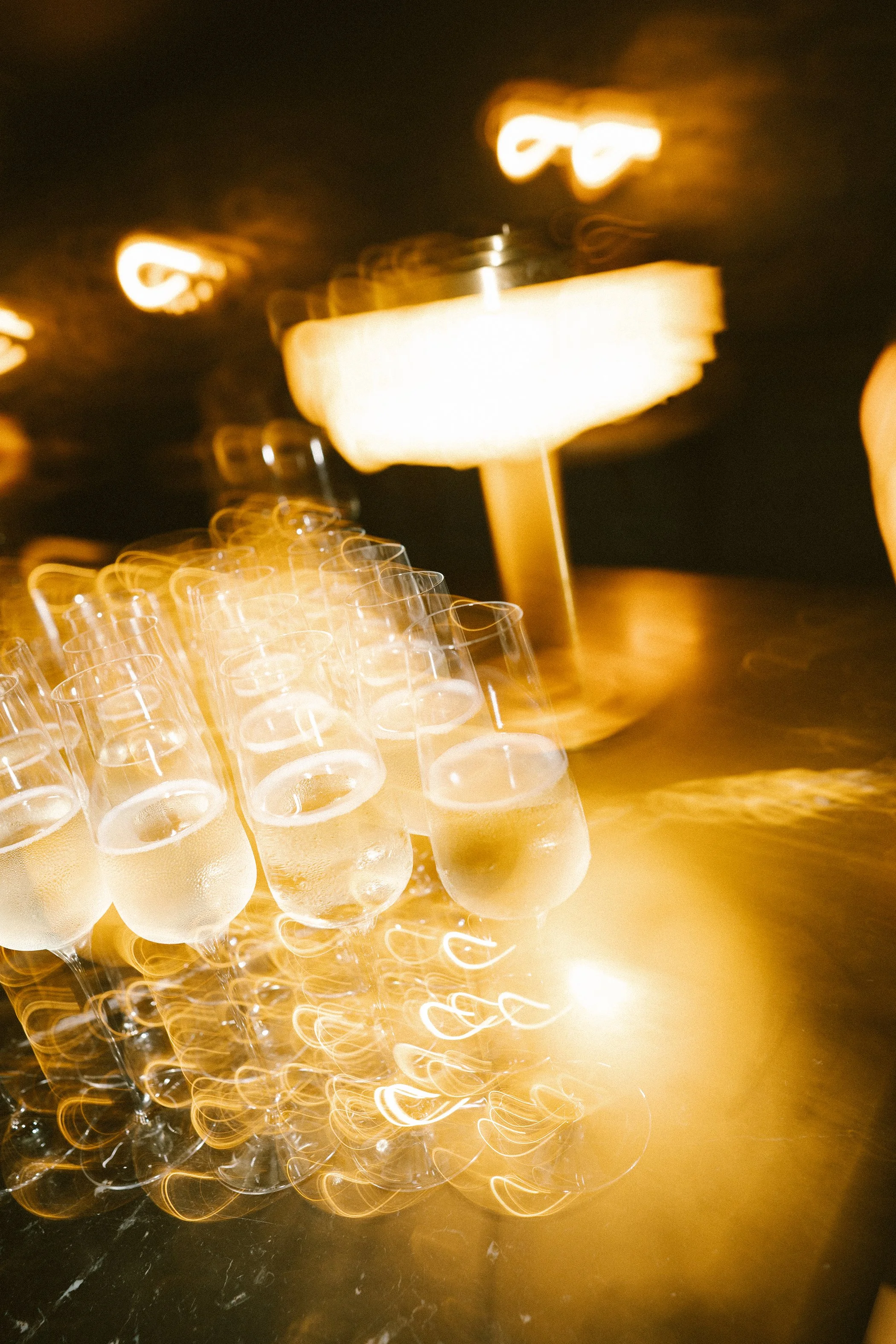 Blurred image of several champagne glasses with condensation, and a central illuminated rectangular light fixture, creating a luminous and abstract scene.